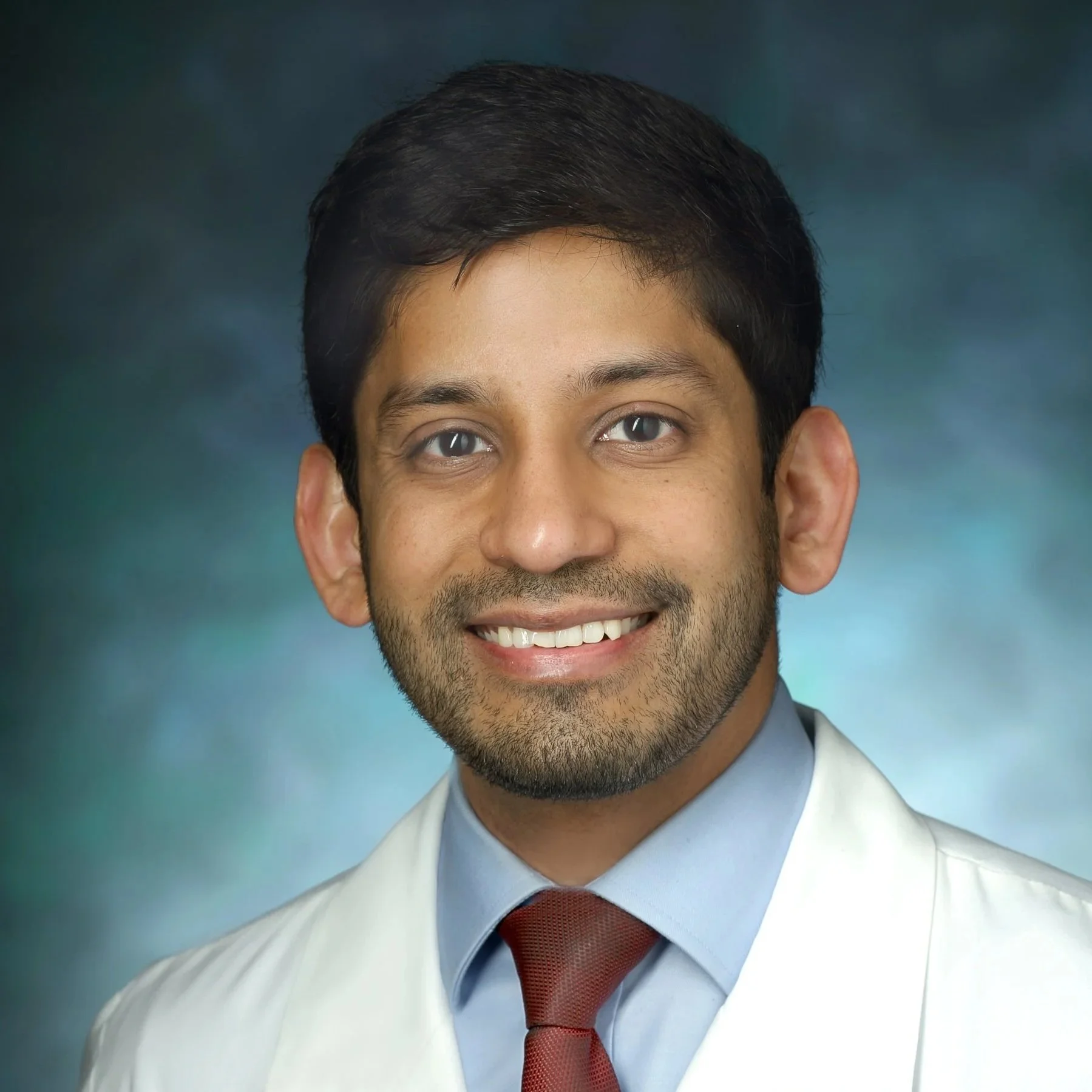 Close-up portrait of a smiling man wearing a white lab coat, light blue shirt, and red tie, with brown hair and brown eyes, against a blue-green background.