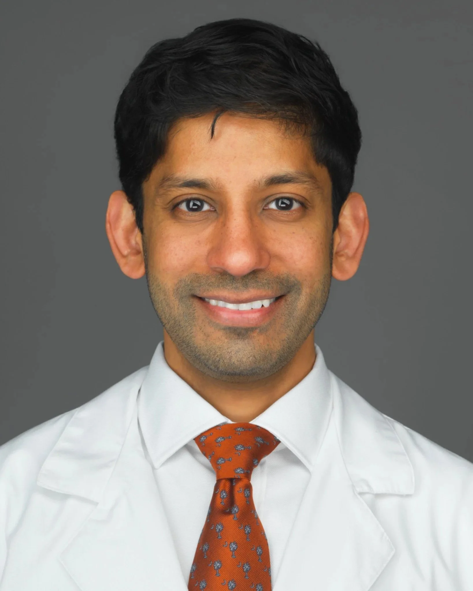Headshot of a man with dark hair, wearing a white shirt and an orange tie with a pattern of small silver trees, smiling against a gray background.