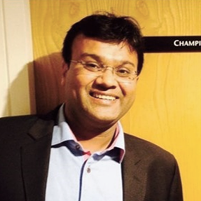 A man with glasses, wearing a dark suit and light blue shirt, smiling in front of a yellow background.