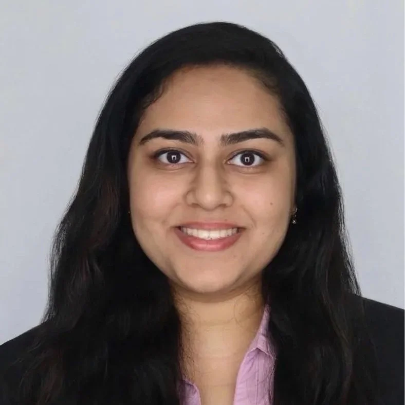 A woman with long black hair, smiling, wearing a black blazer and pink shirt, standing against a plain light gray background.