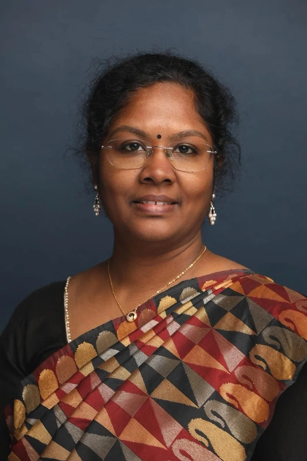 A woman with dark hair, glasses, and earrings wearing a traditional saree against a dark background.
