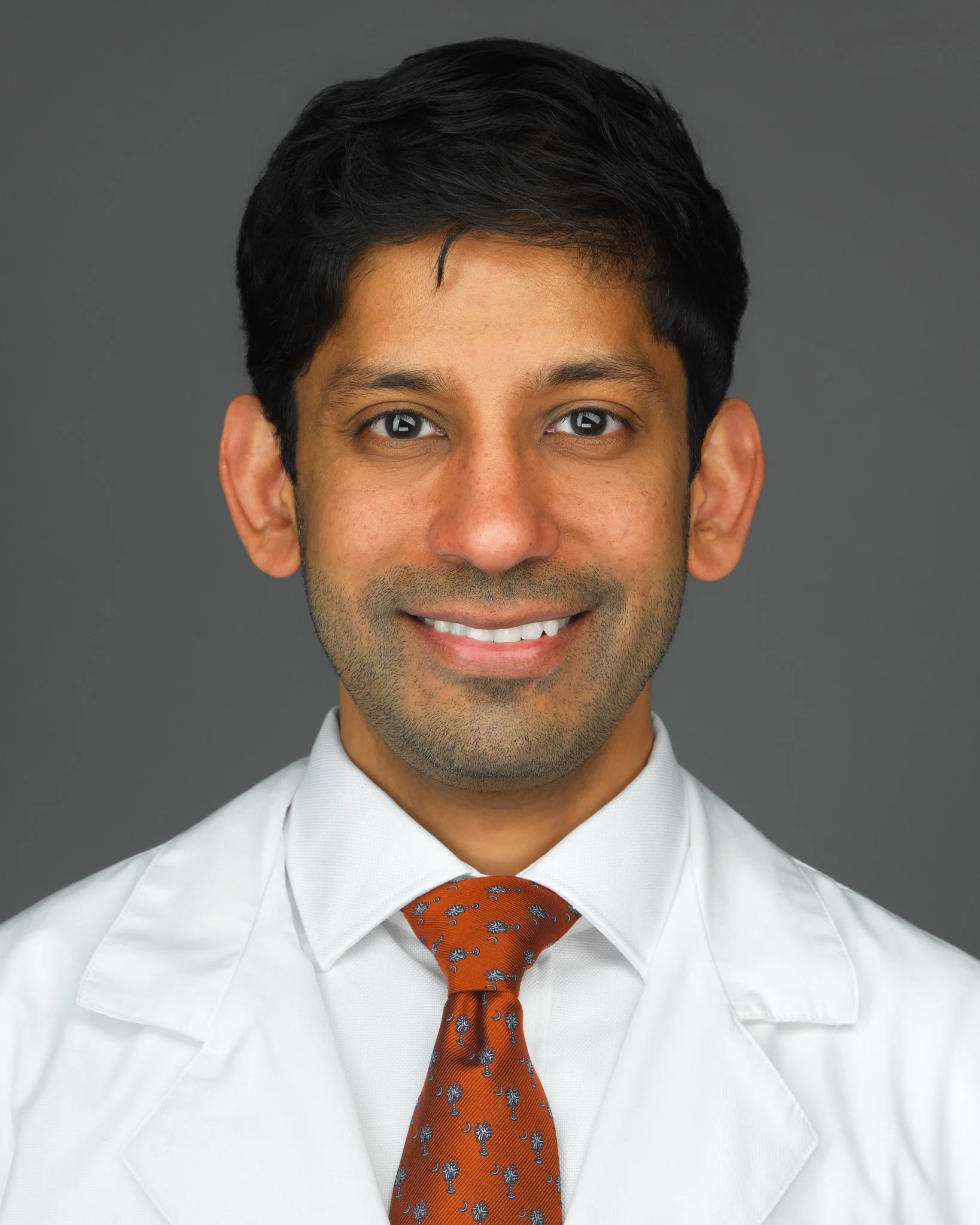 Close-up of a man wearing a white dress shirt, orange tie with palm tree pattern, and a gray background.
