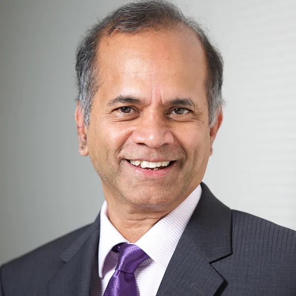 A smiling middle-aged man with dark hair graying at the temples, dressed in a dark suit, white shirt, and purple tie, standing indoors near a light-colored wall.