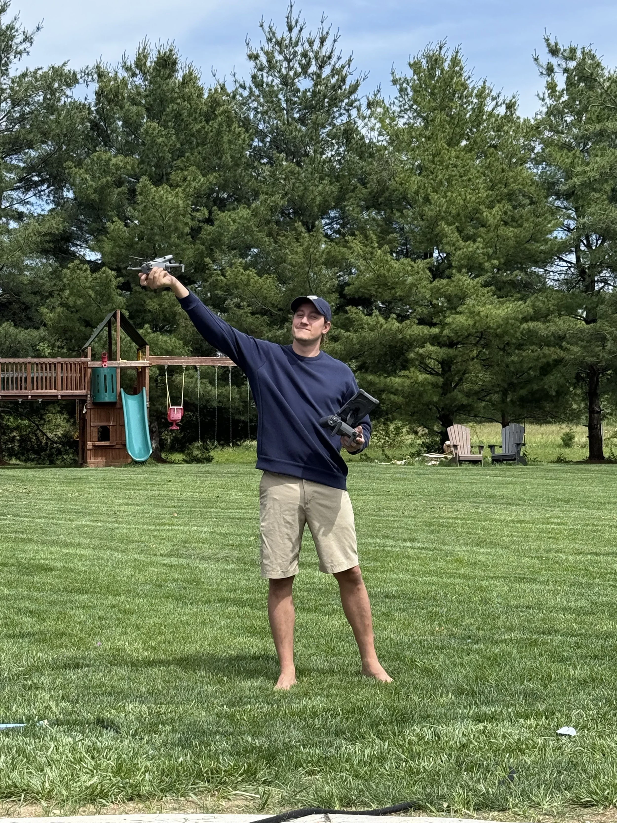 A young man in a blue long sleeve shirt, khaki shorts, and a baseball cap stands barefoot on a grassy lawn, holding a drone in his right hand and a remote controller in his left hand, with a smile. There is a backyard play structure with a slide and swings, some lawn chairs, and tall trees in the background.