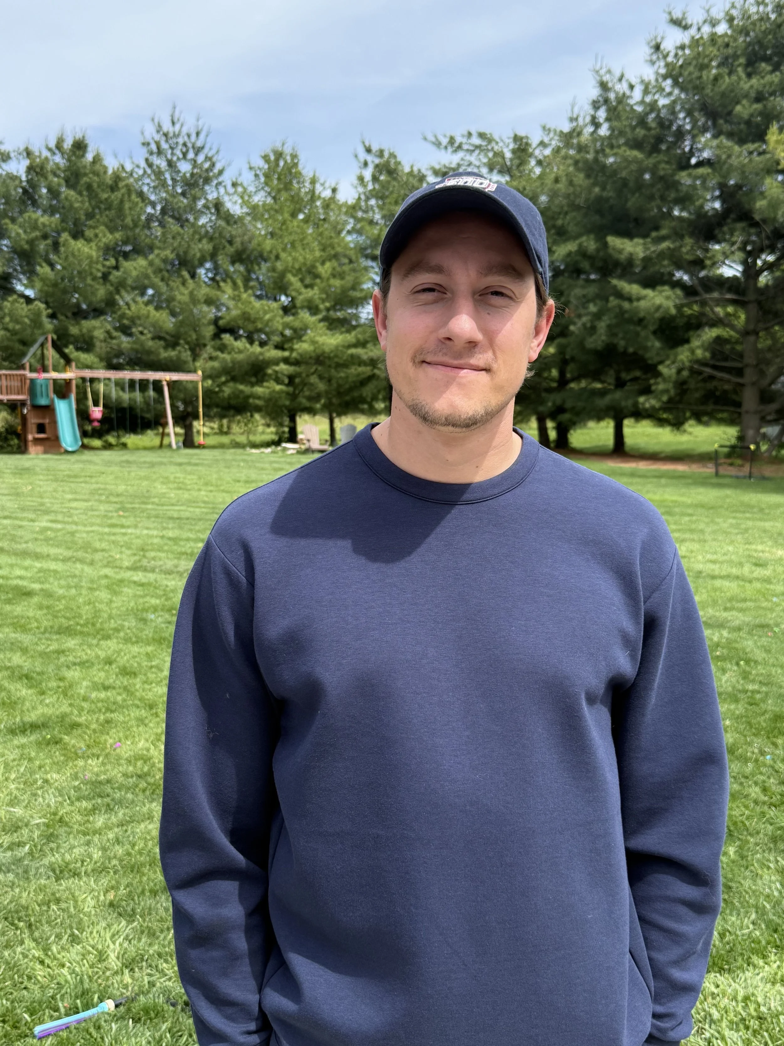 A young man with light skin, short brown hair, and a slight beard wearing a navy blue cap and navy blue long-sleeve shirt, standing outdoors on a grassy area with trees and a play set in the background, under a partly cloudy sky.