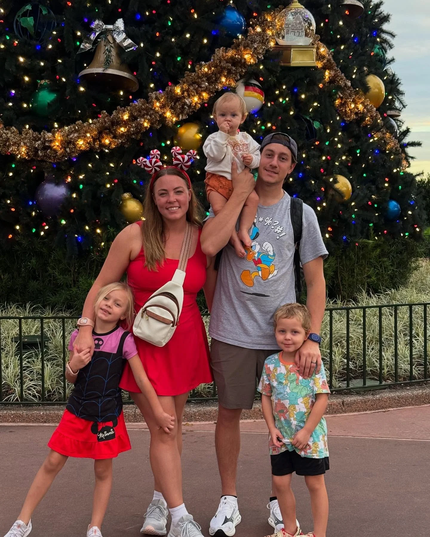 A family of six standing in front of a large decorated Christmas tree with colorful ornaments and lights. The family includes two adults and four children, with the adults smiling and holding the children. The children appear to be ages 3-8, and the background shows an outdoor setting with grass and a fence.
