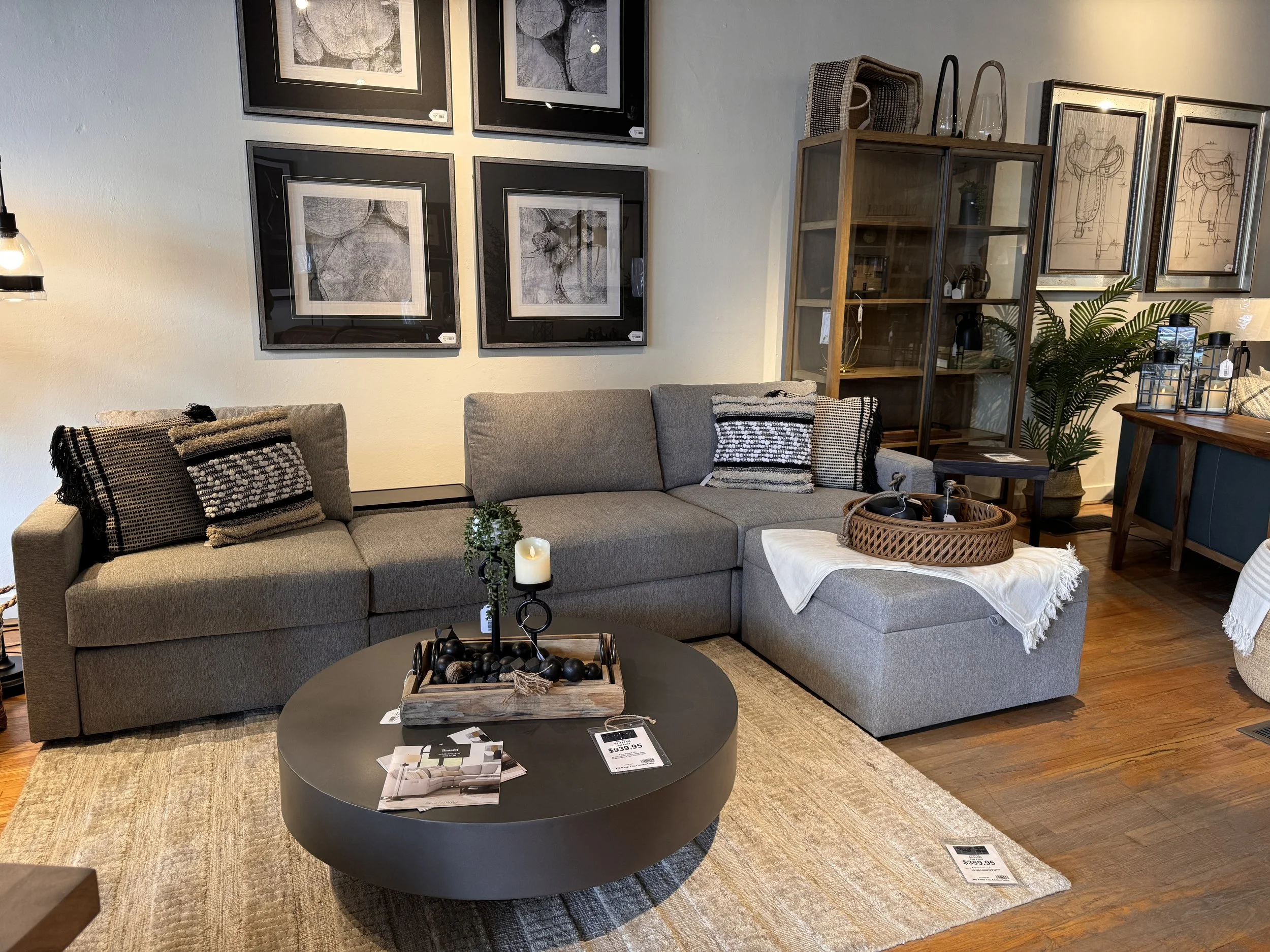 A living room with a gray sectional sofa, black and white patterned throw pillows, a round black coffee table with decorative items, wall art, a glass-front wooden cabinet, and a wooden side table with houseplants.
