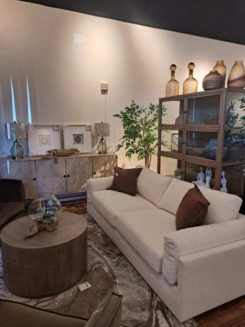 Living room with beige sofa, dark brown pillows, wooden coffee table with a glass jar, sideboard with framed art, lamps, vases, and a large green plant.