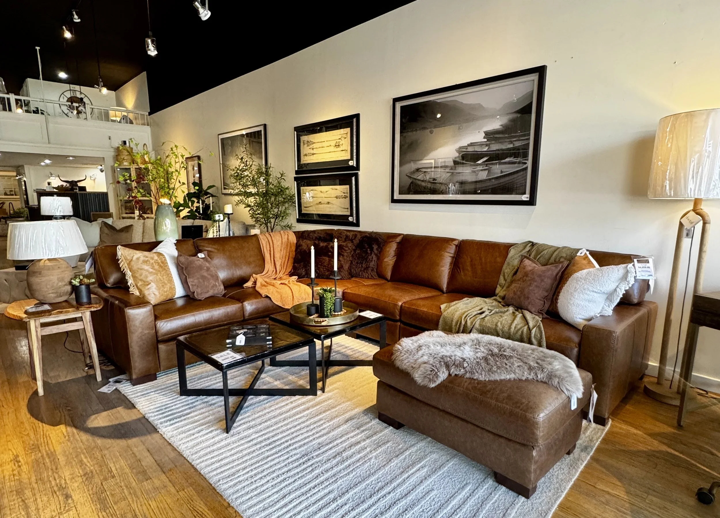 A cozy living room with a large brown leather sectional sofa decorated with various pillows and throws, a rectangular black coffee table, a beige area rug, and two standing lamps. There are framed black-and-white photographs and artwork on the wall, and various houseplants and decorative items on the side tables and in the background.