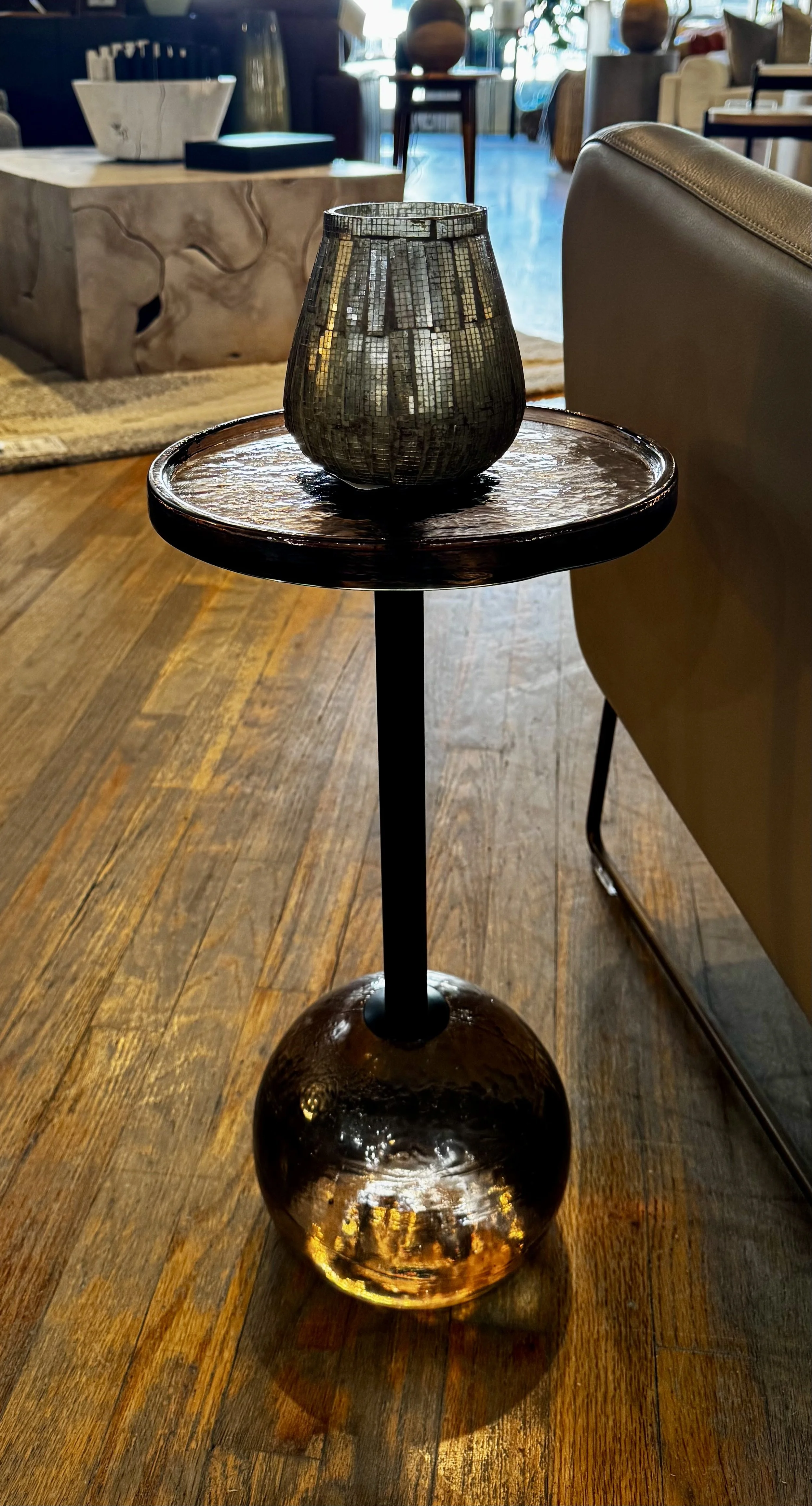 Decorative mosaic vase on a small round side table with a dark amber glass round top and spherical amber-colored base, in a cozy interior setting with wooden flooring and modern furniture.