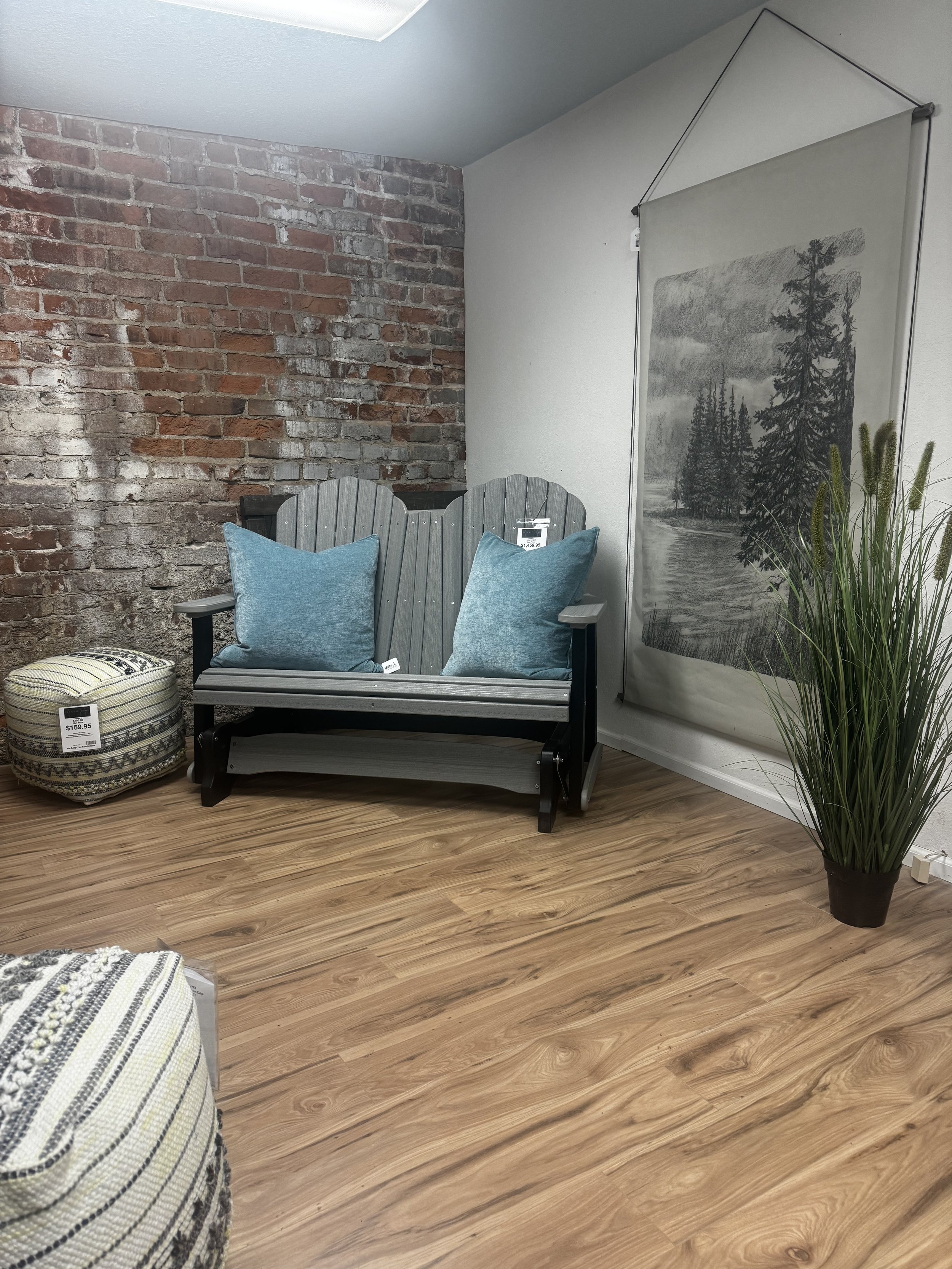 A small decor display featuring a gray wooden gliding loveseat with two blue pillows, a patterned pouf, a tall potted grass plant, a tapestry of a black and white nature scene, set against a brick accent wall and wooden floor.