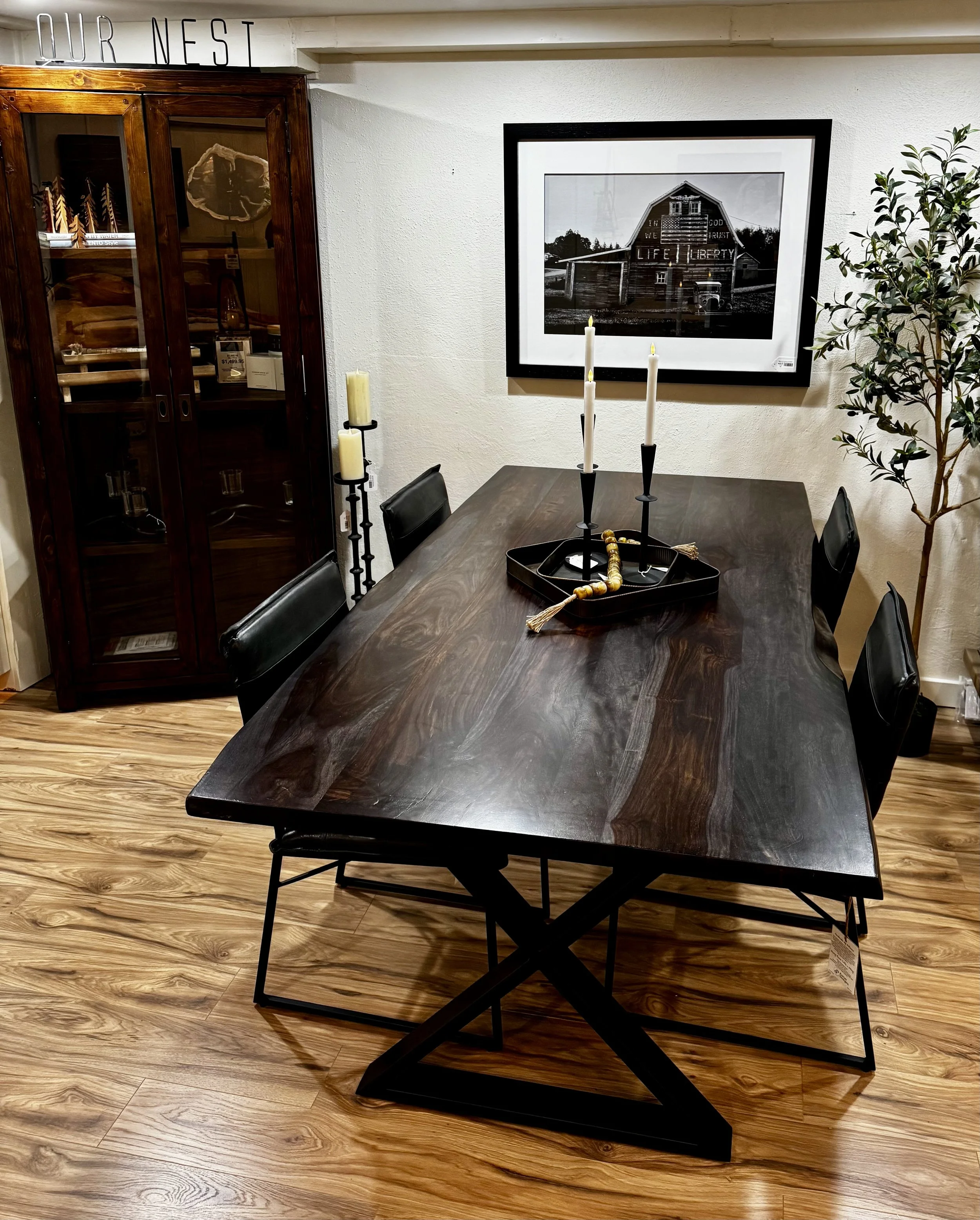 A wooden live edge dining table with black leather chairs, candle holders with candles, and a decorative tray with beads, in a room with a cased wooden cabinet and a framed black-and-white photo on the wall.