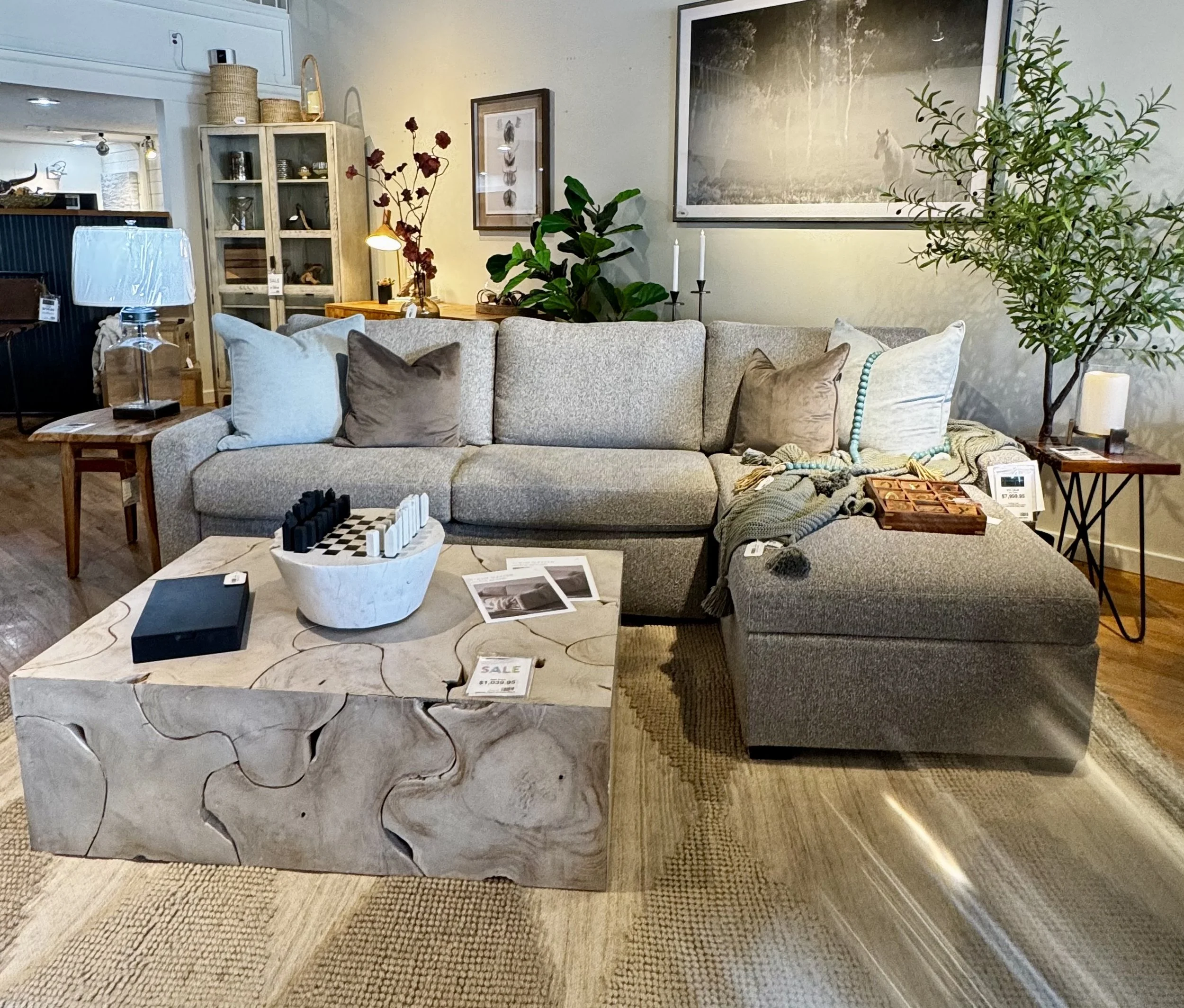 Living room with a gray sectional sofa, puzzle piece teak coffee table, and various decorative items including pillows, plants, and framed pictures on the wall.