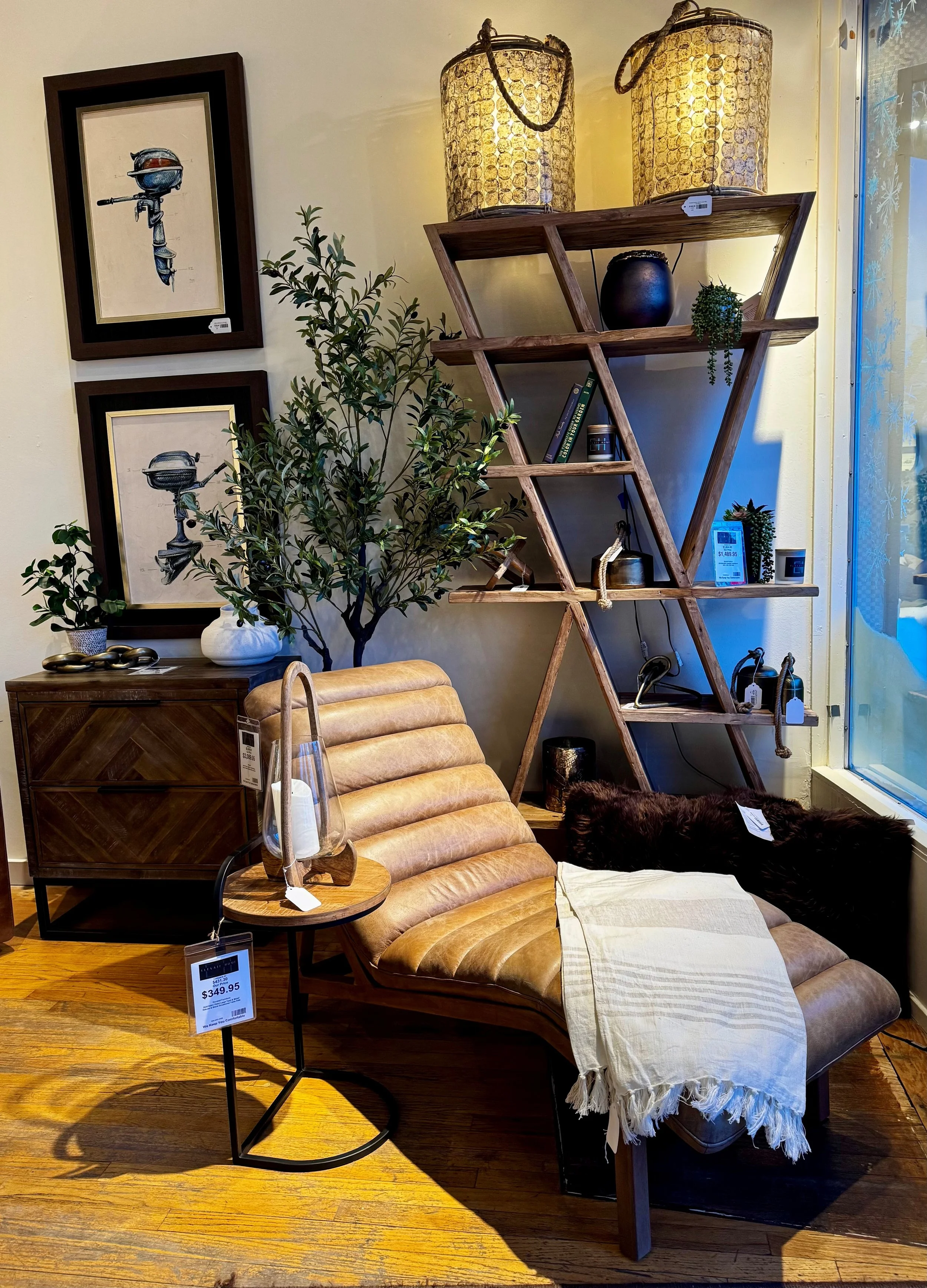 A retail display with a tan leather lounge chair, a small wooden side table with a glass lantern and candle, a wooden display shelf with decorative objects, plants, and framed artwork of a water scooter, all in a cozy, modern style.