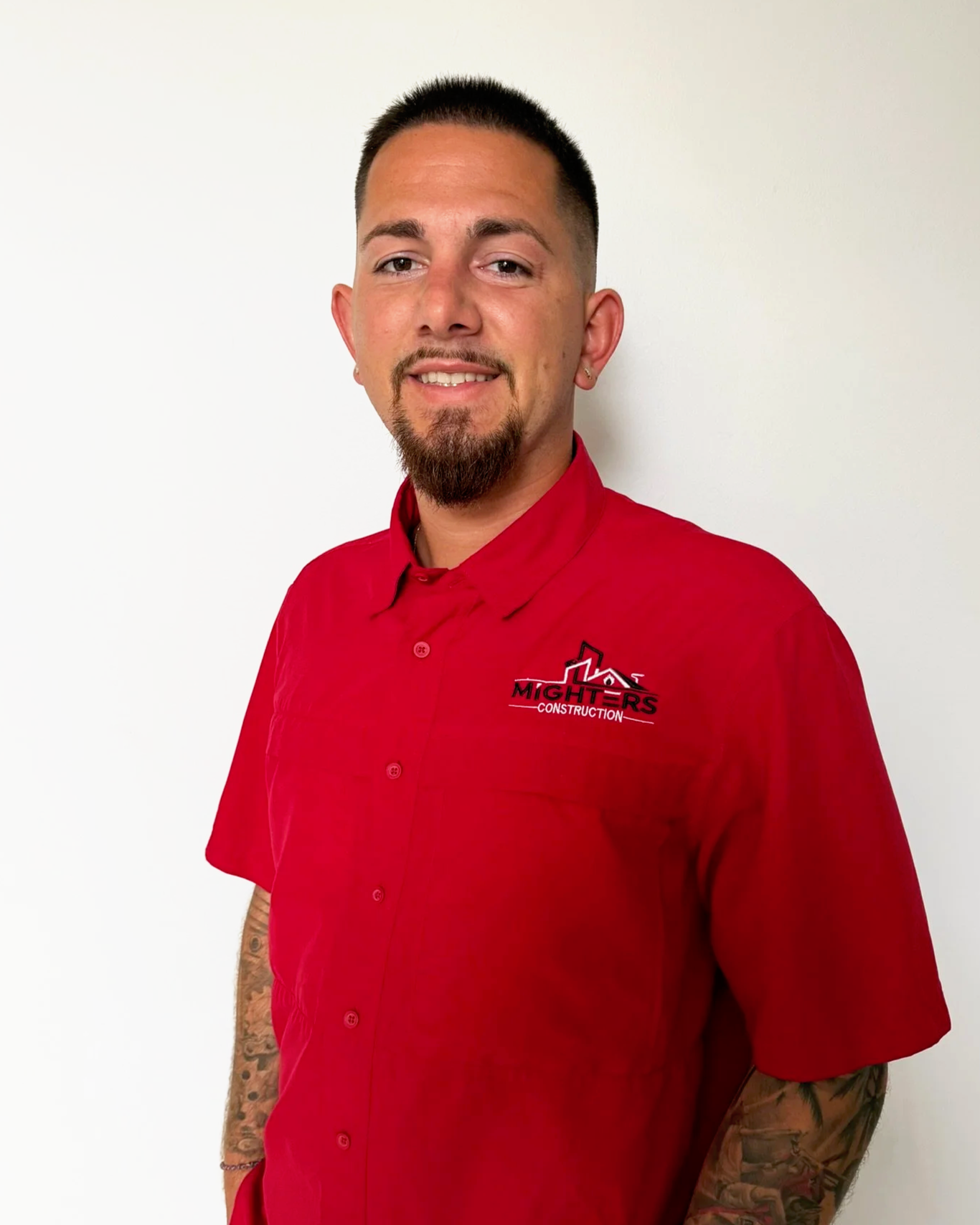A man dressed in a red work shirt with the logo 'Mighters Construction' on it, standing against a plain white background, smiling slightly with a goatee and tattoos on his arm.