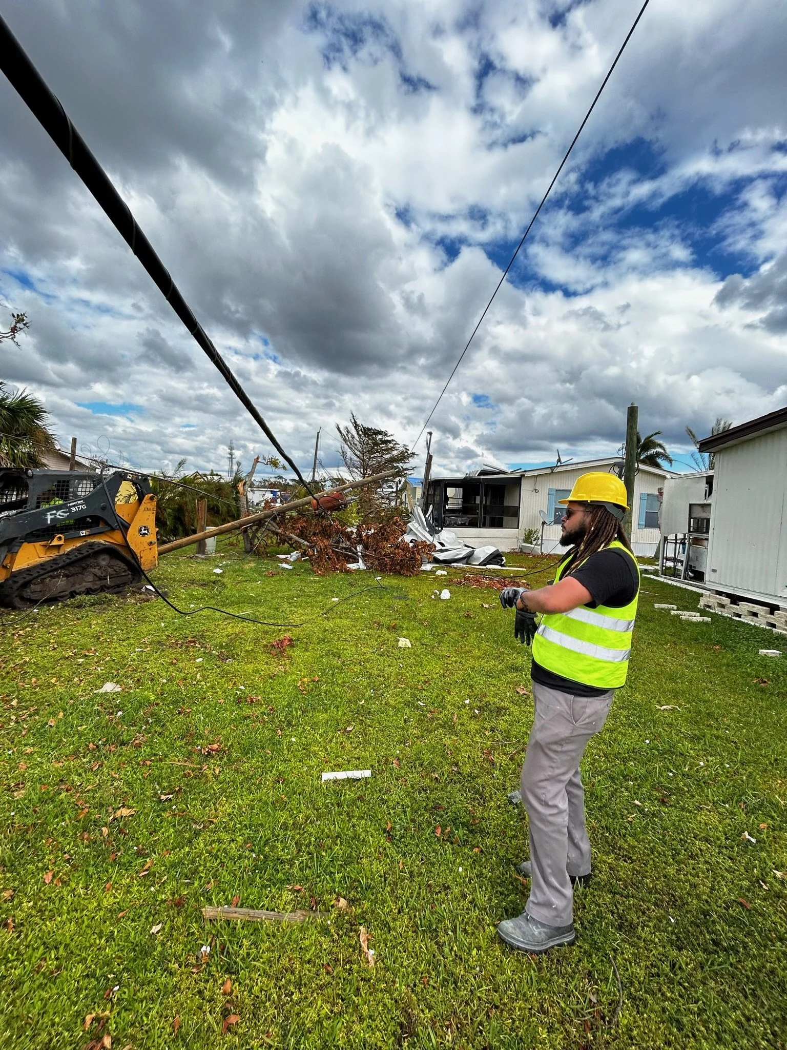 A man wearing a yellow safety helmet and a bright yellow vest stands on a grassy area with debris and fallen trees after a storm. A large fallen tree and a damaged building are visible in the background, along with dark storm clouds in the sky.