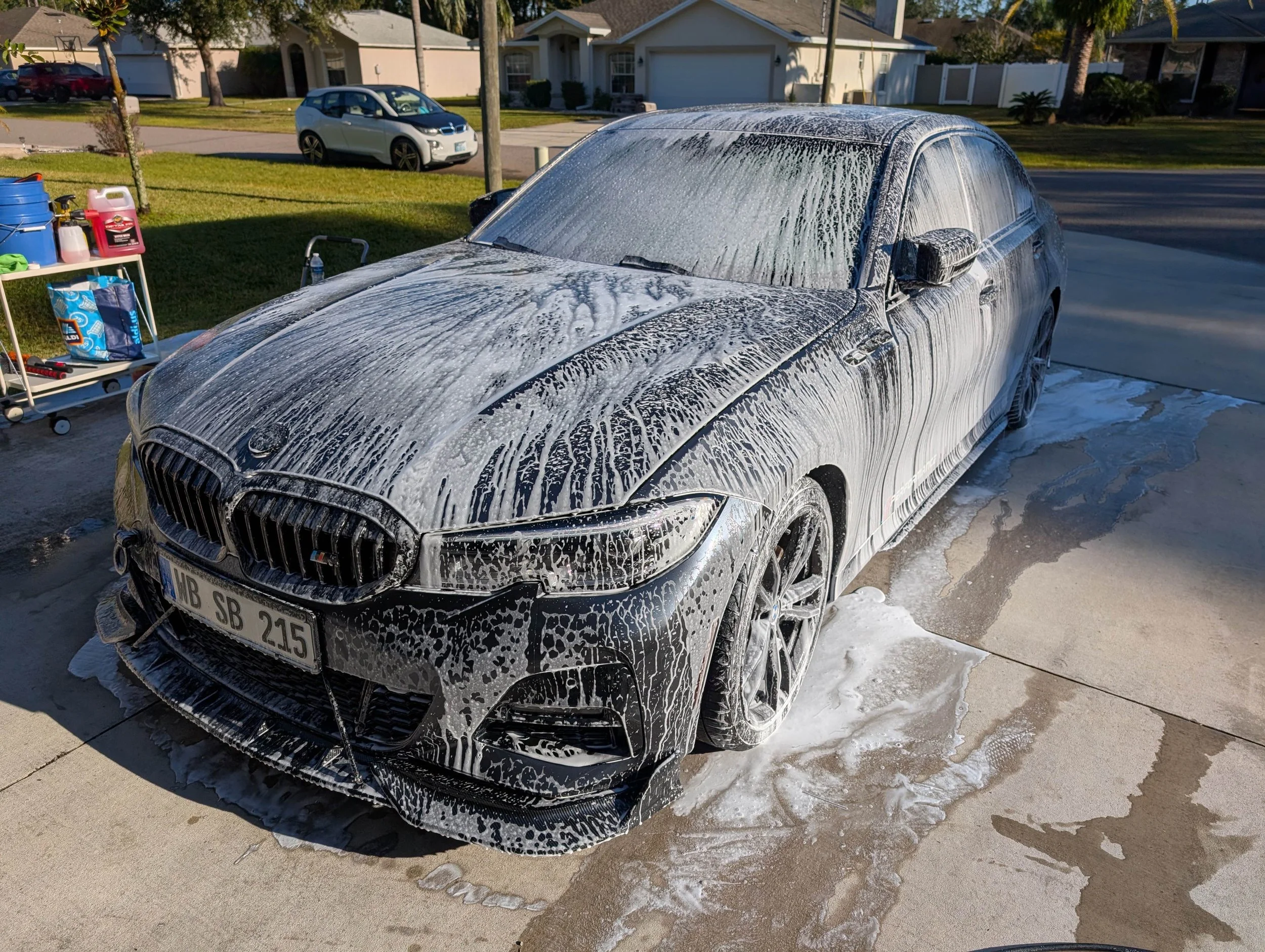 Snita Brookman's BMW is being washed, covered in soap suds, in a residential driveway during the daytime. There are trees, houses, and another car in the background.