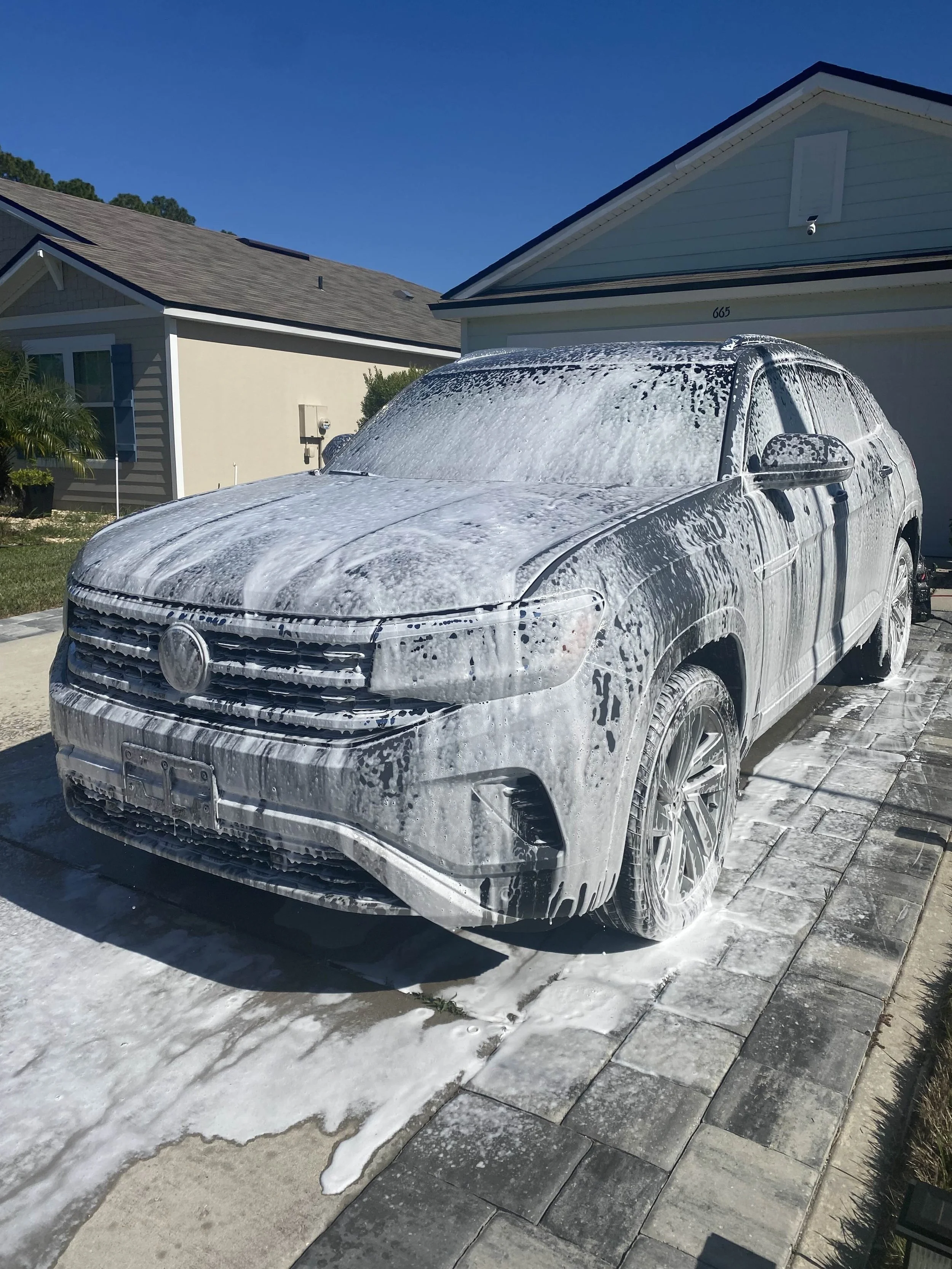 A gray Volkswagen SUV covered in soap suds during a car wash in a driveway with a house in the background and a clear blue sky.