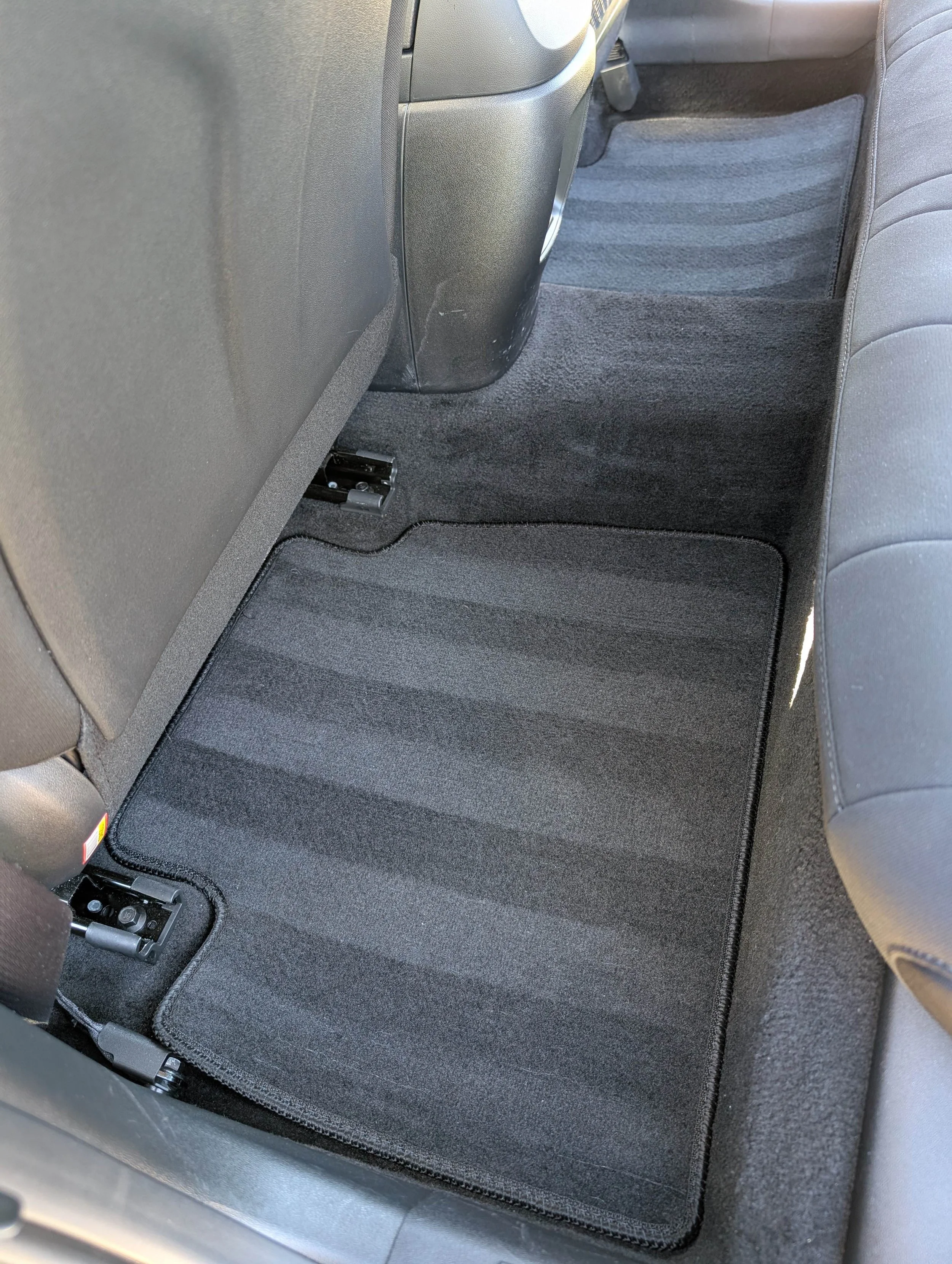 The back seat area of a car showing the black floor mat, the rear of the driver's seat, and parts of the front passenger seat.