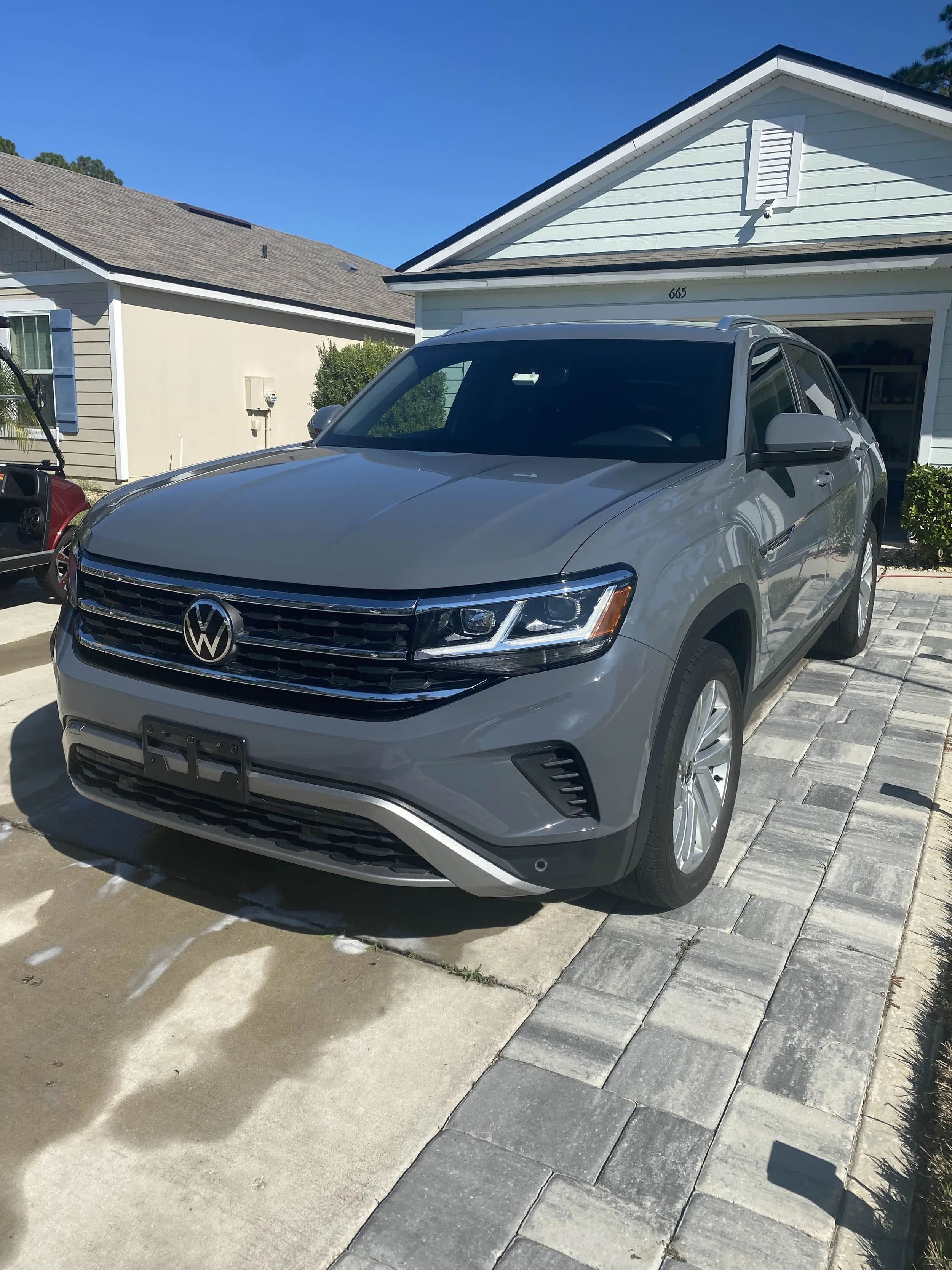 A silver Volkswagen SUV parked in a residential driveway in front of a house with white siding. The house has a garage with the door open, and the sky is clear and blue.