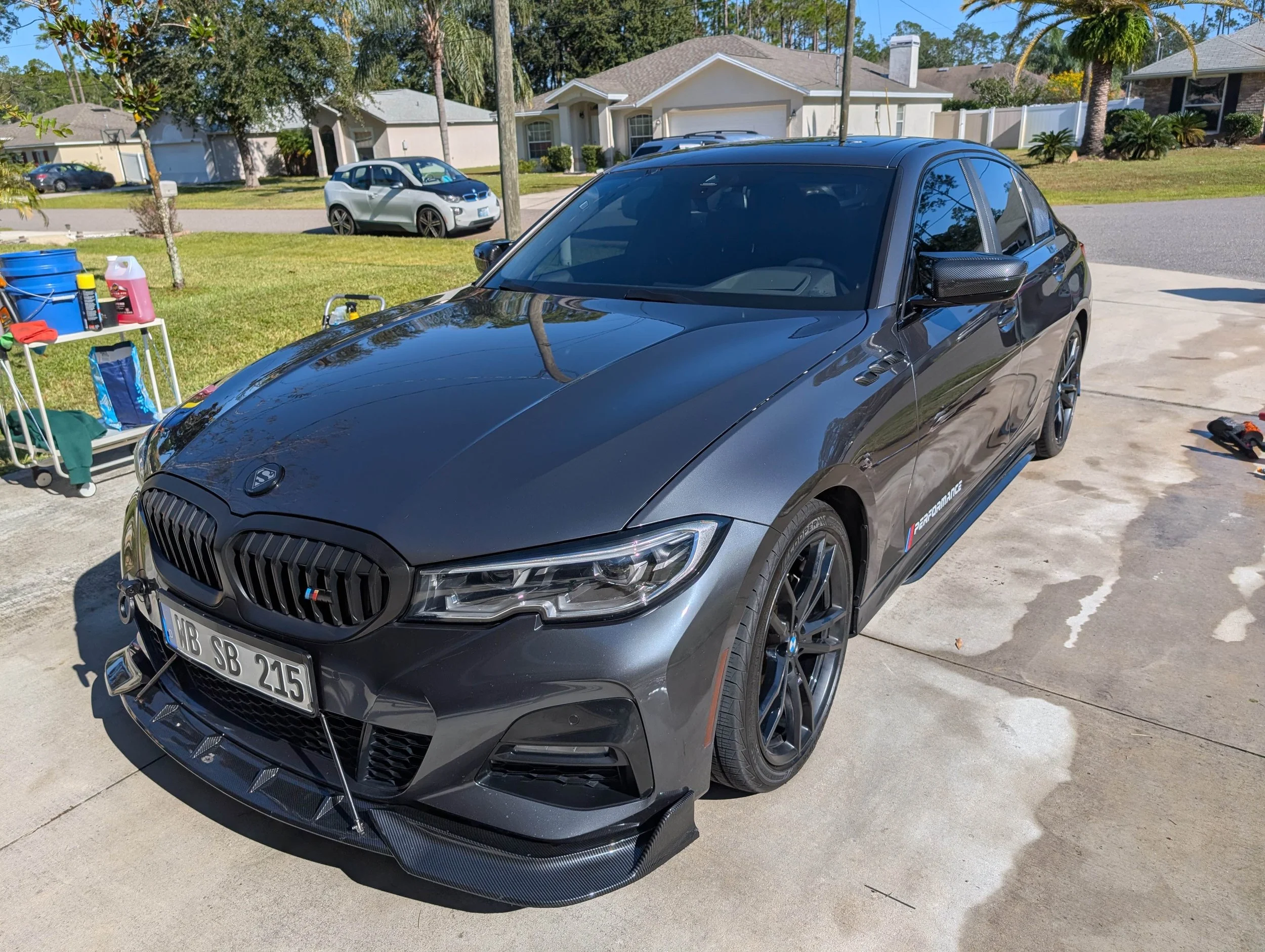 A black sports car, possibly a BMW M model, parked in a driveway with a reflection of a palm tree on its hood. There is a white shelf on the left with cleaning supplies, and a small tool on the ground nearby. In the background, a silver electric car and a residential neighborhood with houses, trees, and a clear sky are visible.