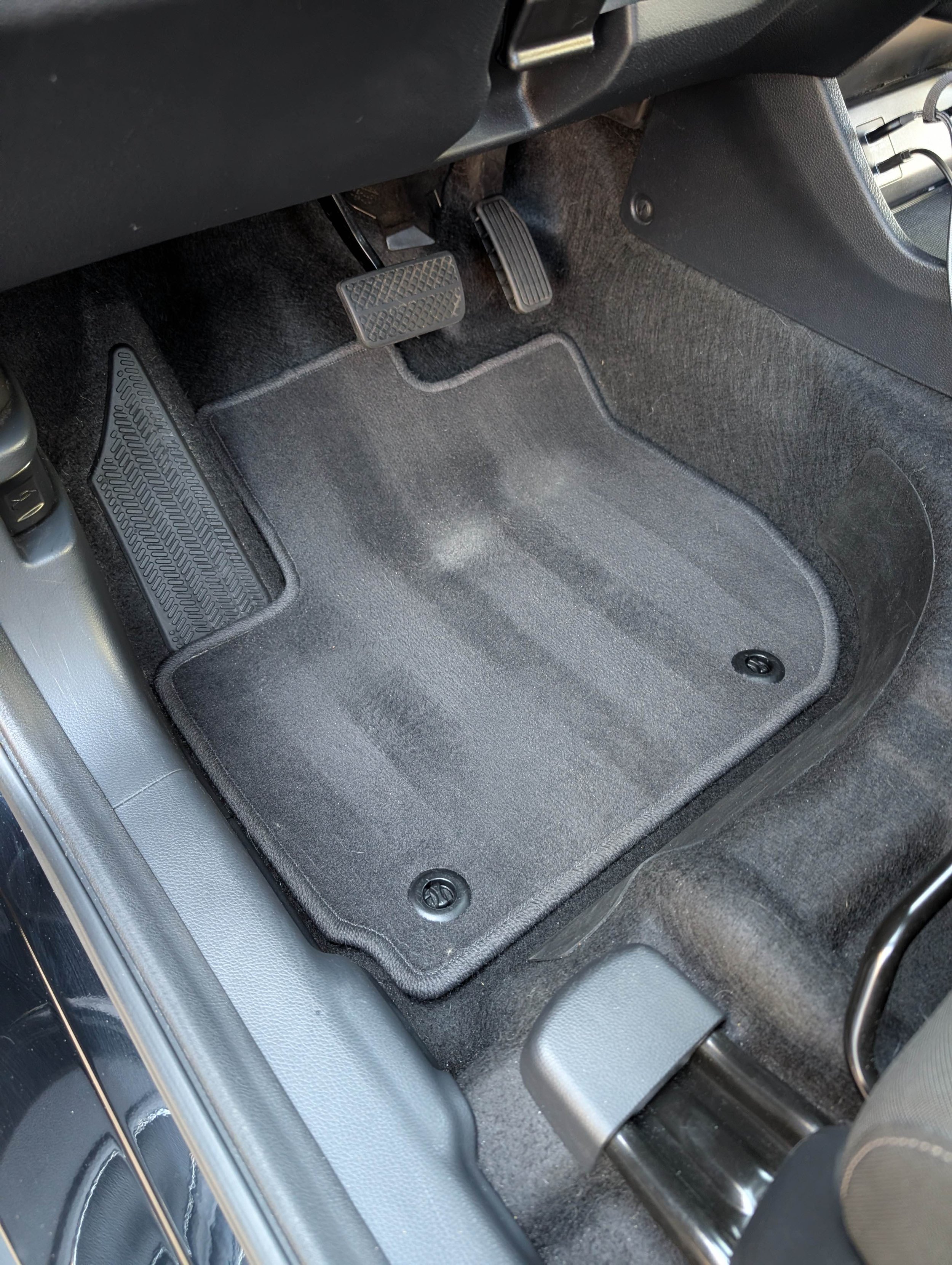 The image shows the inside of a vehicle near the driver’s side footwell area, including the accelerator pedal, brake pedal, and a rubber floor mat.