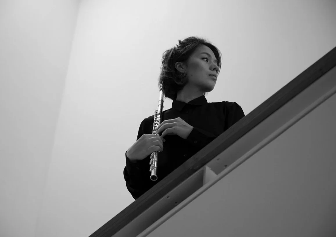 A black and white photo of a woman holding a flute, looking to the side, standing beside a staircase railing.
