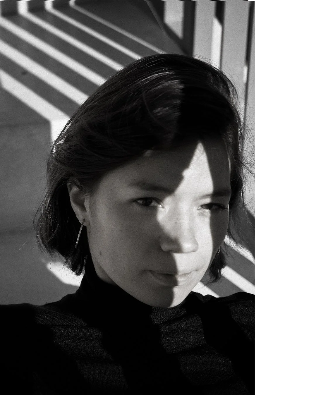 A black and white close-up of a woman with dark hair, wearing a dark top and earring, taking a selfie with sunlight casting a shadow across her face.