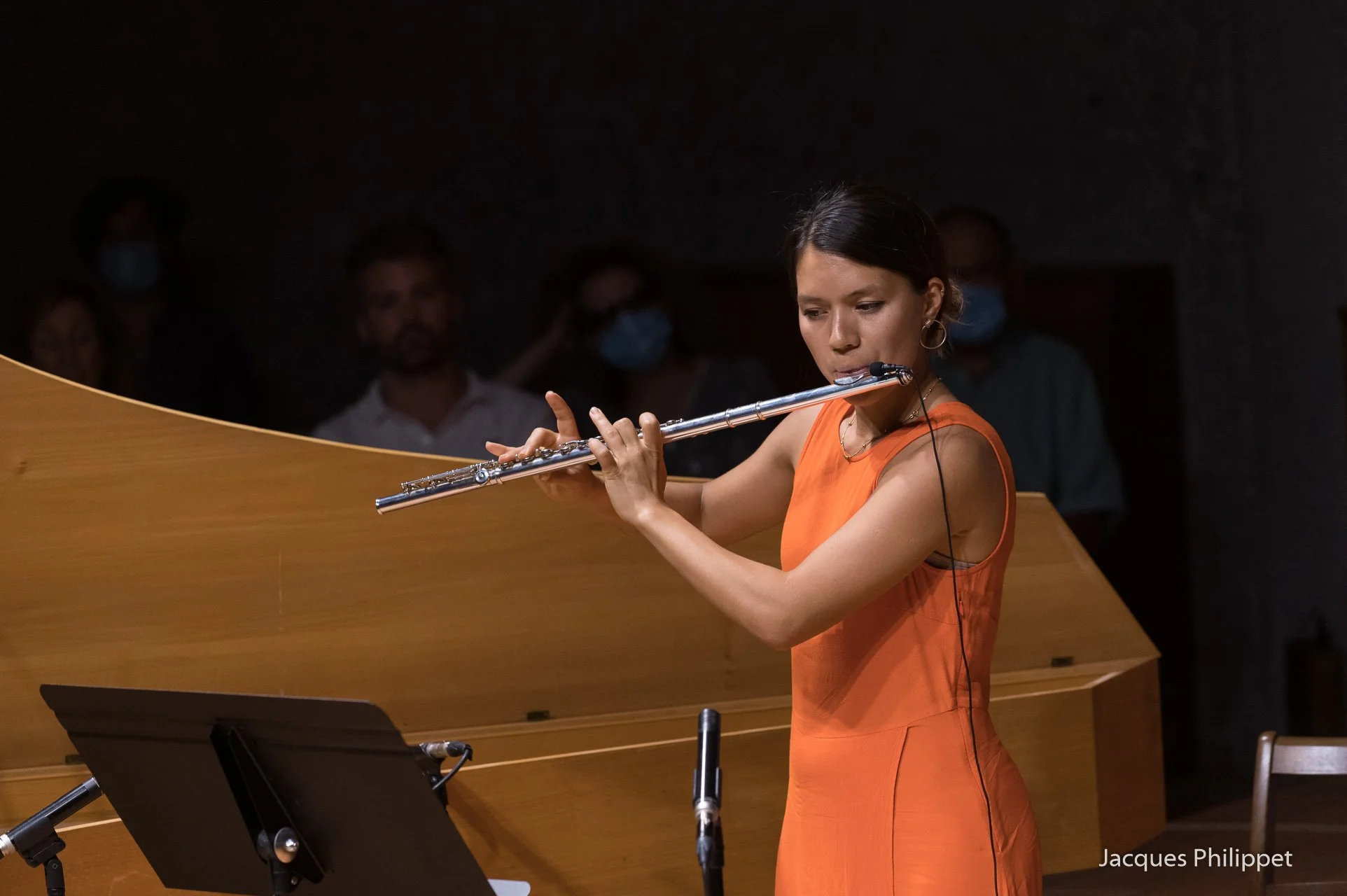A woman in an orange dress playing a flute on stage during a performance, with an audience in the background.