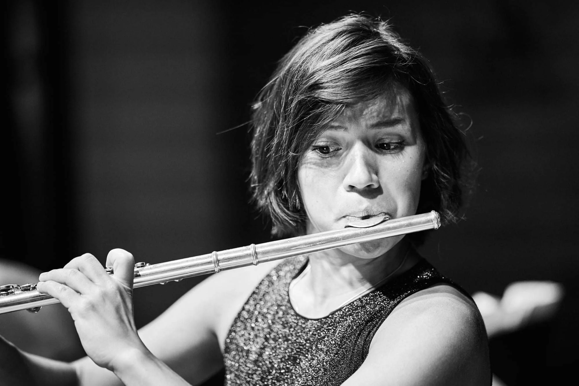 Woman playing a flute, with short hair, wearing a sleeveless, sparkly top, captured in black and white.