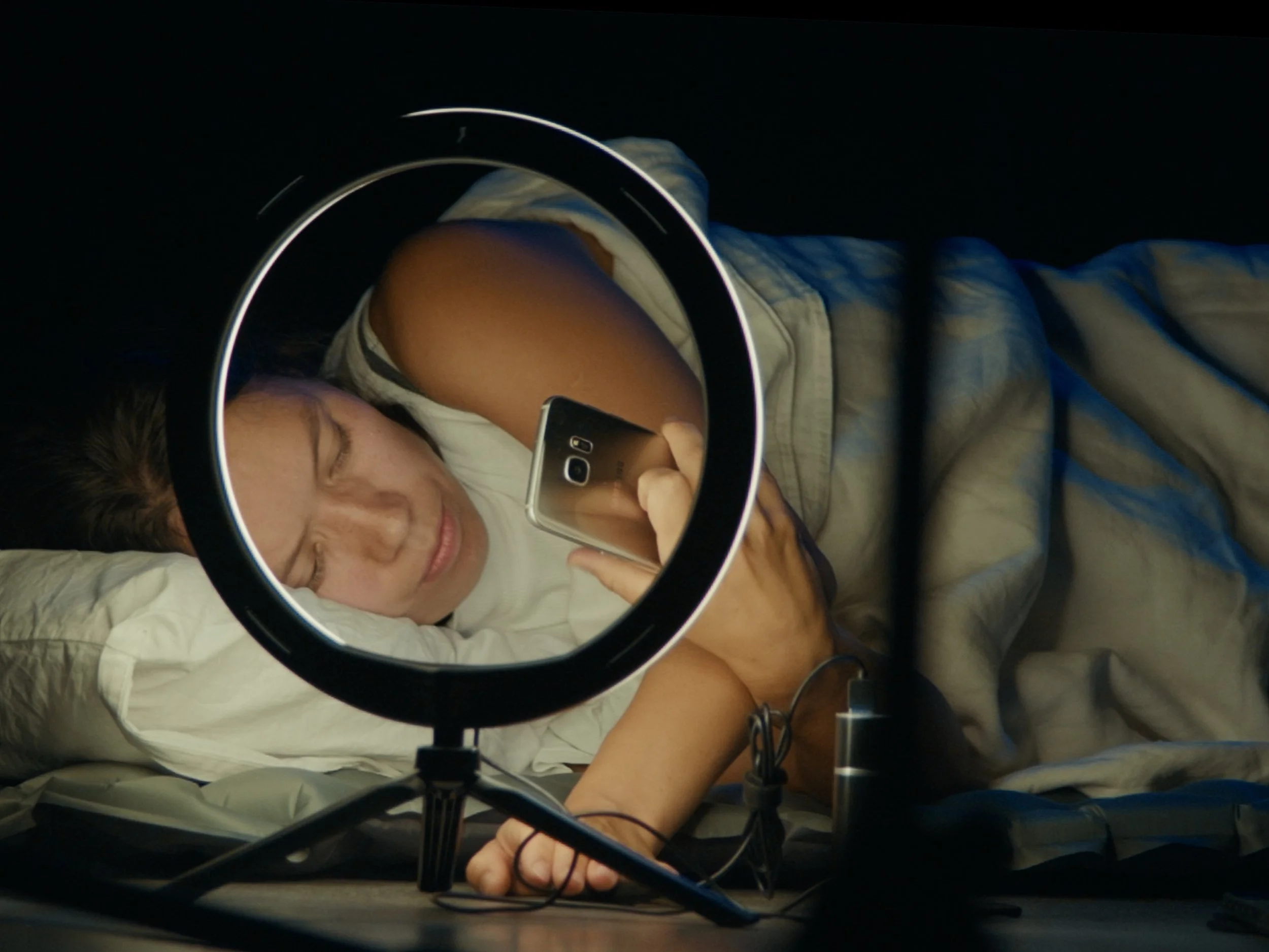 A person lying in bed taking a selfie in a ring light, with a smartphone in hand. The person is wearing a sleeveless shirt and has closed eyes, with a pillow behind their head.