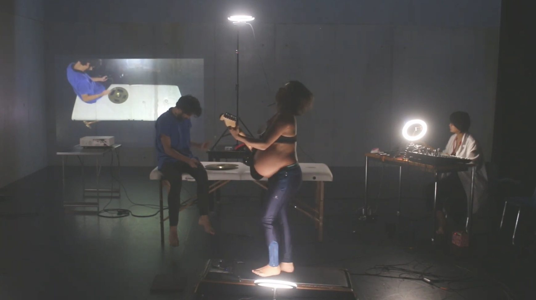 A woman in a bra and jeans playing an electric guitar while a man sits on a chair with a metal object on his lap, a woman in a white coat at a table with a ring light, and a projection of an image of a person with a metal object on a white surface on a wall.
