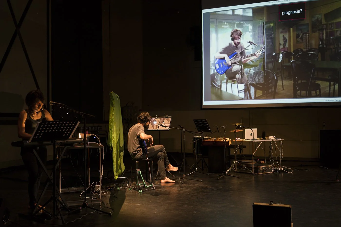 Musicians performing on stage with electronic equipment and a large screen displaying a photo of a man playing guitar in a cafe or restaurant.