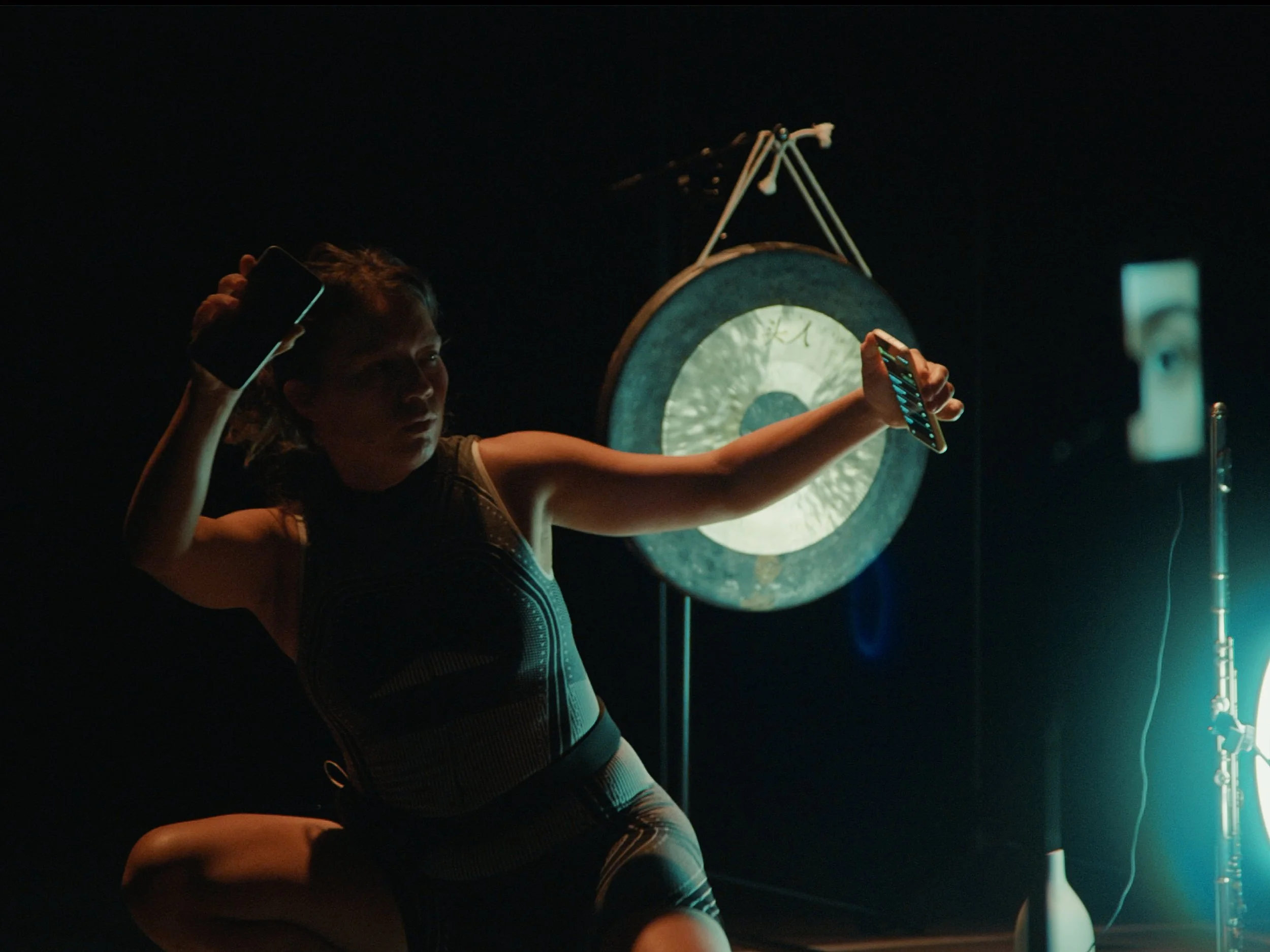 A woman is taking a selfie in front of a large gong, with a gong stick and other musical equipment around. The scene is dark with a glow from a nearby light.