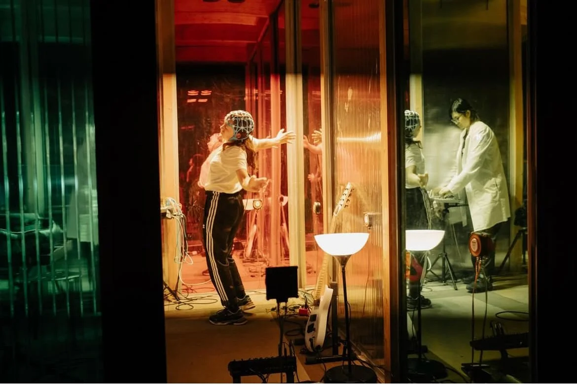 A woman in a white shirt and black pants with white stripes on the sides, wearing headphones, standing in front of a glass wall, gesturing with her hands, with a man in a white lab coat inside a recording studio, playing a keyboard, with music equipment and lighting.