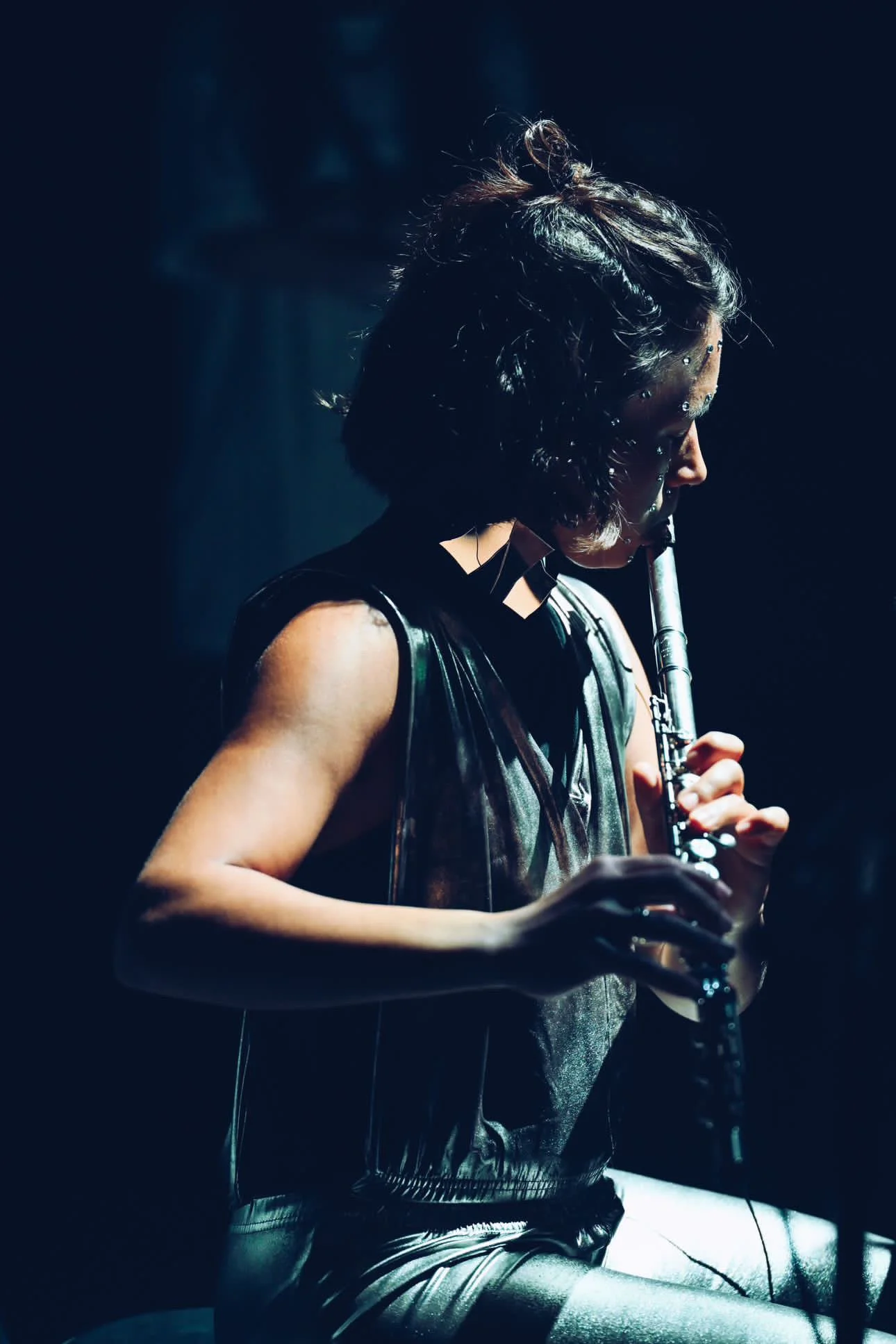 A woman playing a flute in dim lighting, with short curly hair and wearing a dark, shiny outfit.
