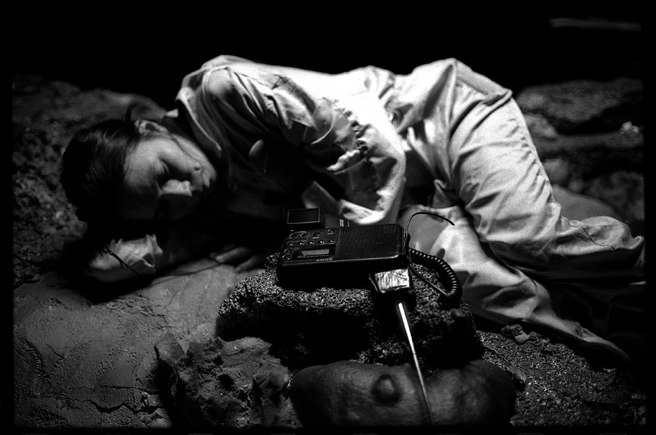 A person lying on the ground in a space suit beside a radio device and a microphone or antenna, in a dark environment.