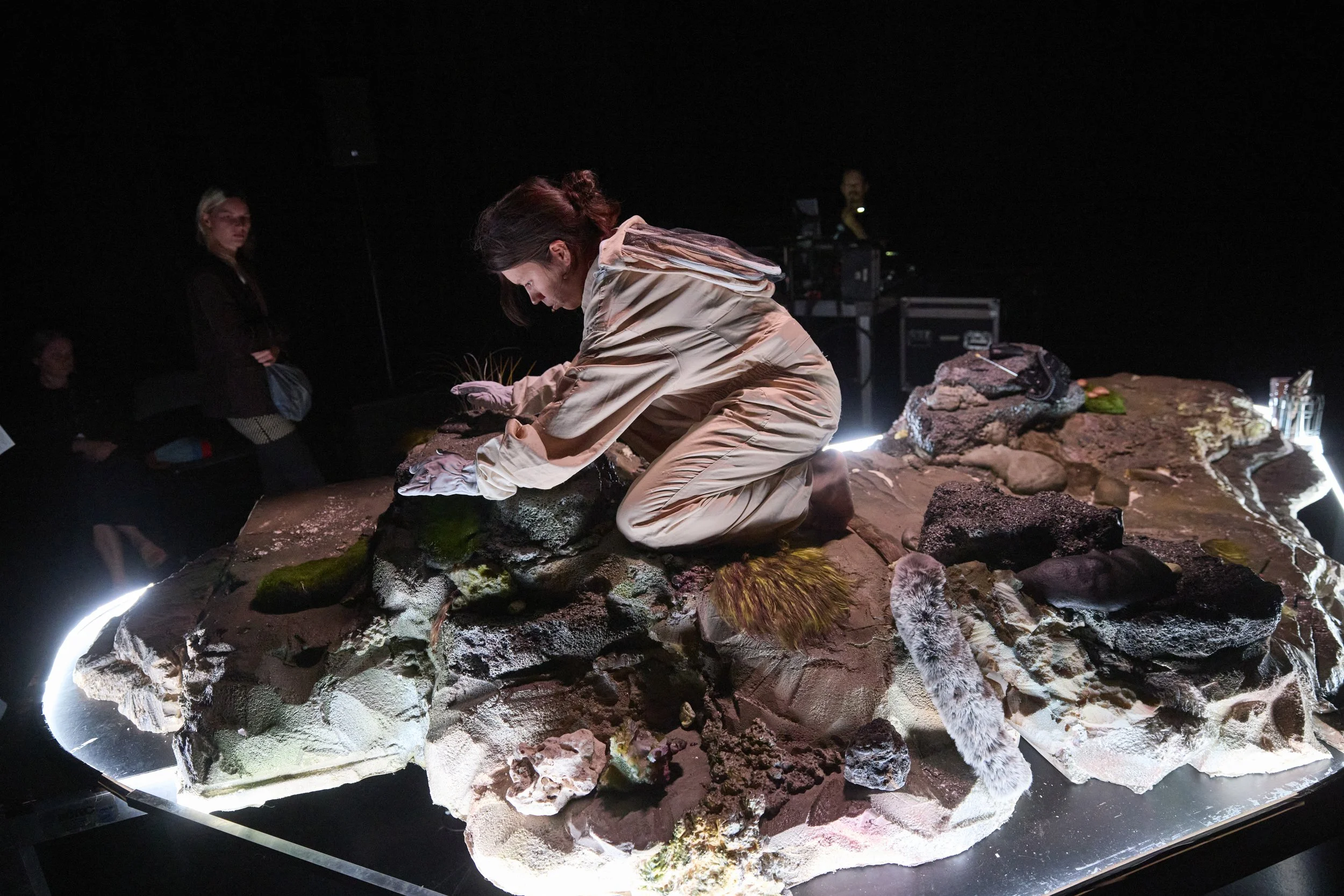Person kneeling on a rocky terrain with dark background, engaged in a detailed activity, surrounded by other people and equipment.