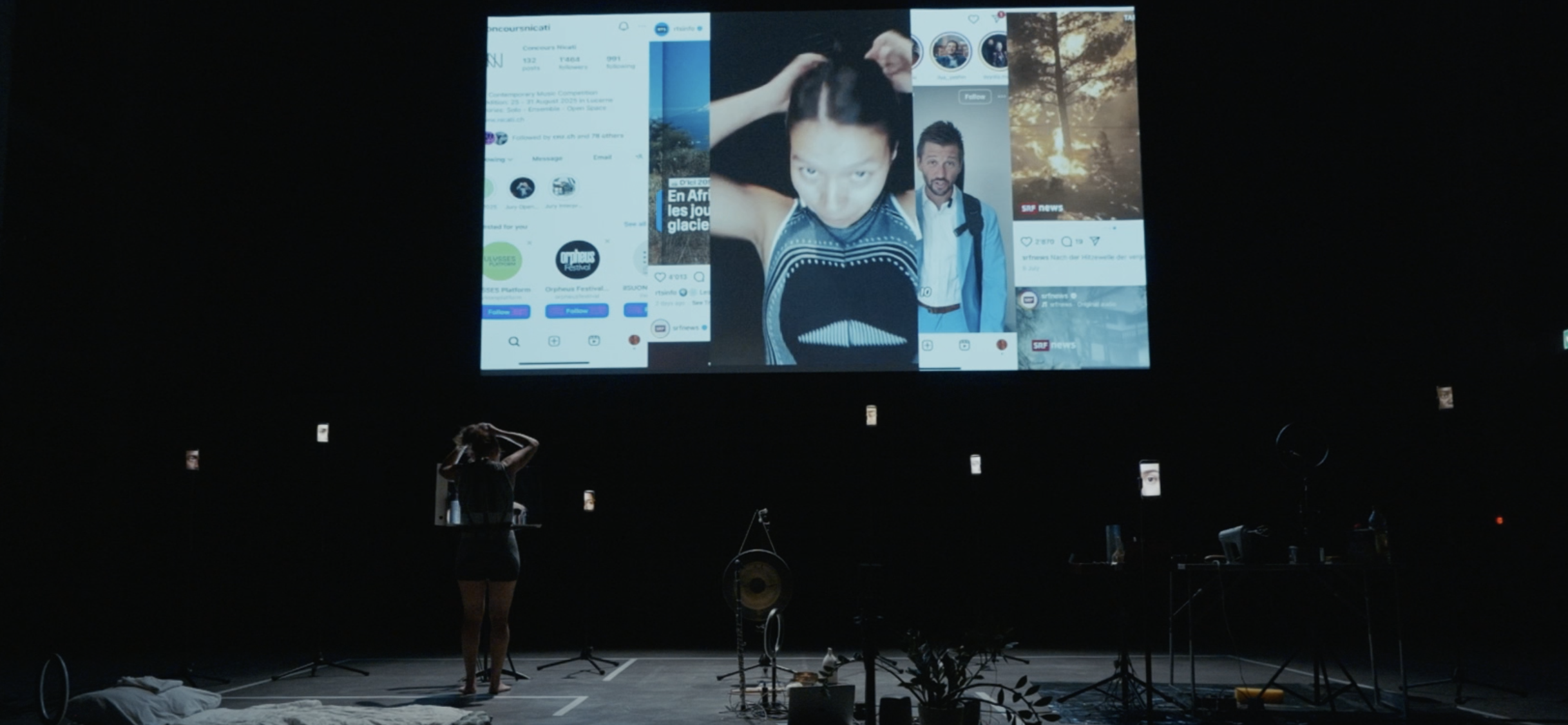 Large indoor stage with a giant screen displaying a collage of smartphone images and a woman in black adjusting her hair in front of music stands and equipment, with dim lighting.