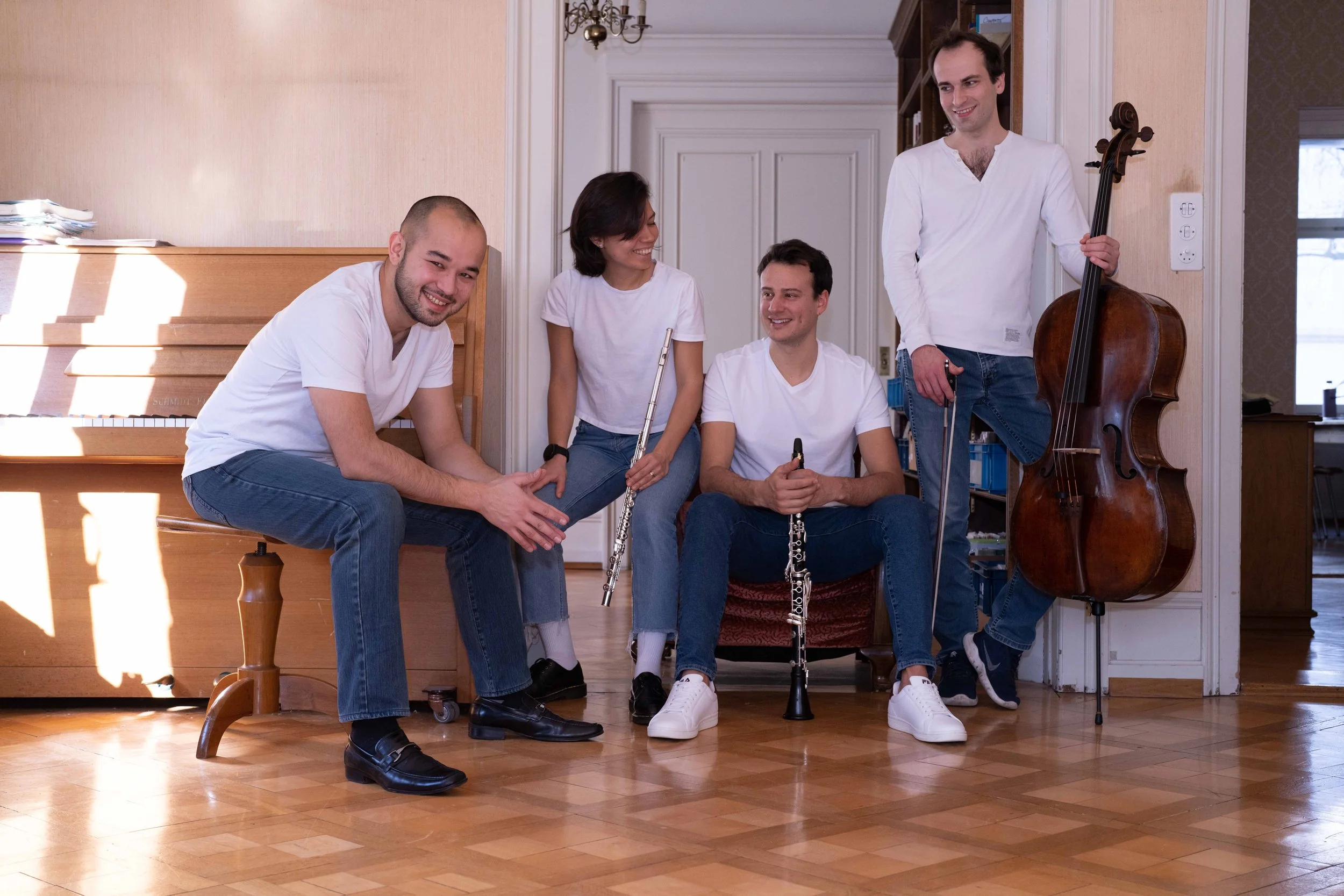 Five people sitting and standing in a room with musical instruments, smiling and chatting. One person plays the piano, two hold flutes, one has a clarinet, and one stands with a double bass.