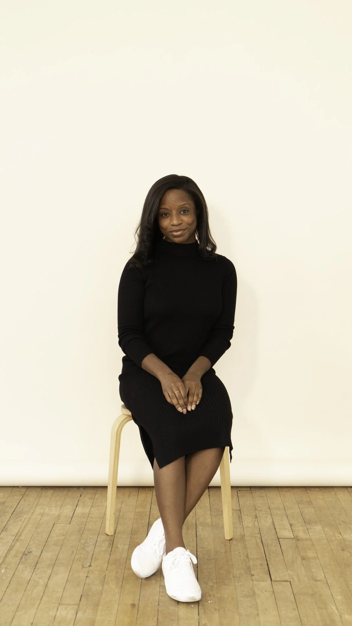 A Black woman sitting on a wooden stool against a plain off-white background, wearing a black long-sleeve ribbed dress, white sneakers, and looking at the camera.