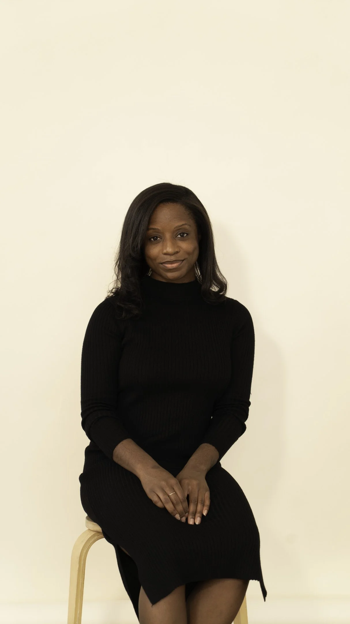A Black woman with dark hair, sitting on a wooden stool against a plain cream-colored wall, wearing a black long-sleeve dress, smiling subtly, with her hands resting on her lap.