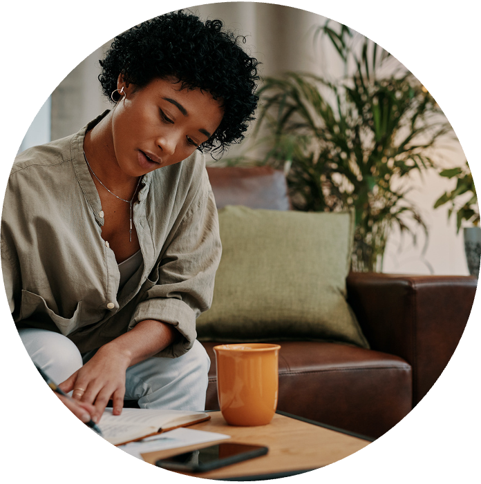A Black woman with short curly black hair, wearing a beige shirt, sitting on a couch and looking down at papers on a coffee table with an orange mug nearby, in a cozy living room with plants in the background.