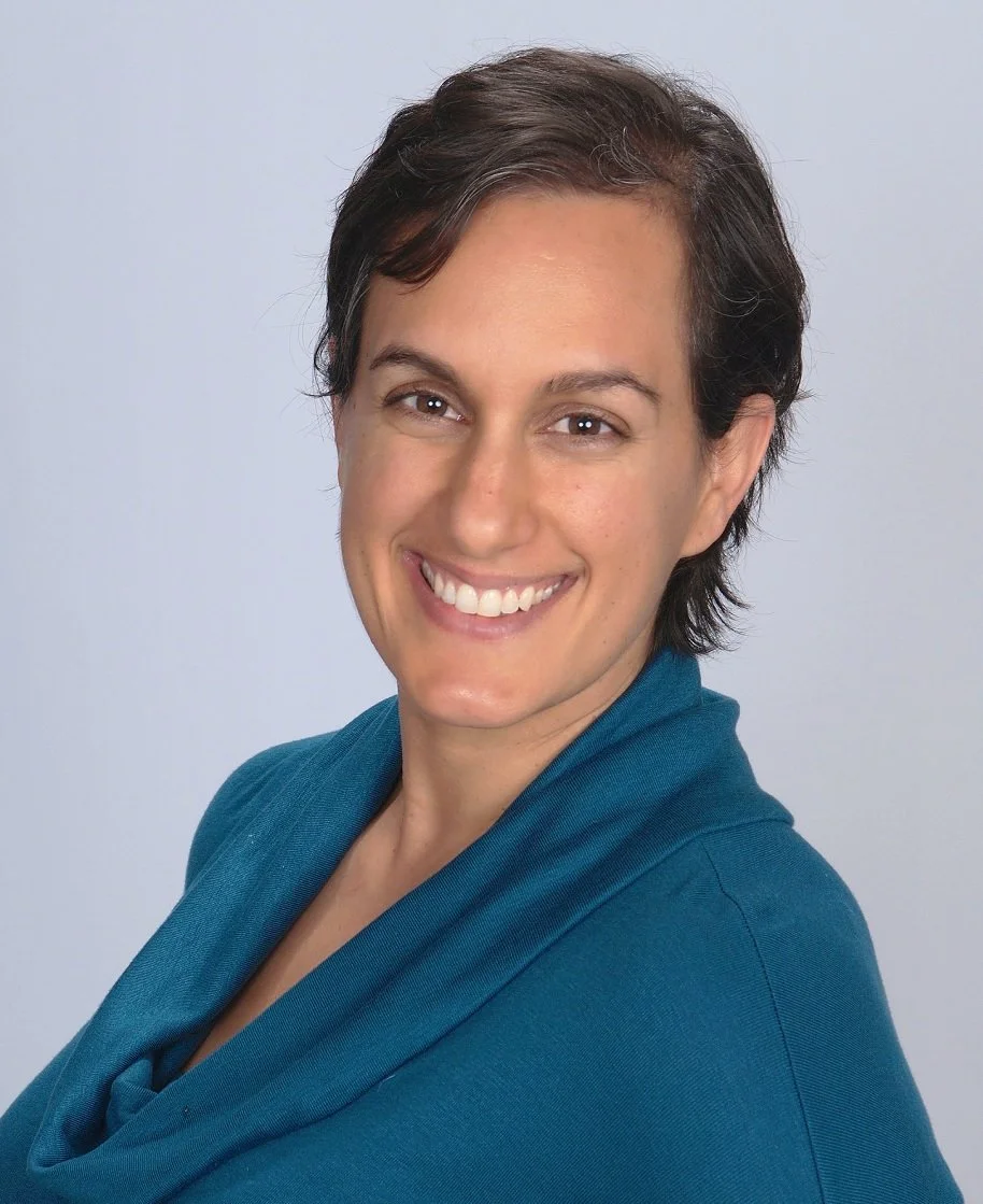 Portrait of a woman with short dark hair, smiling, wearing a teal blazer against a light background.