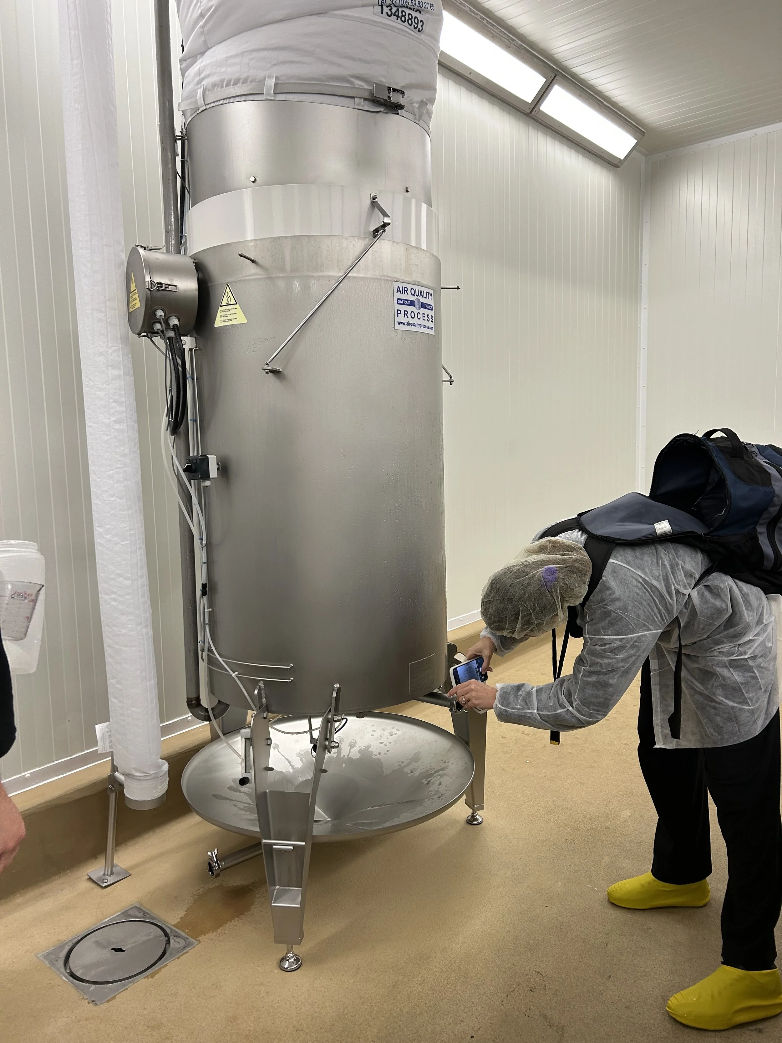 A person wearing yellow protective shoe covers, a gray jacket, and a hairnet is taking a photo of a large stainless steel machine in a clean, white-walled room. The machine is labeled with a sticker that reads 'Air Quality Process.' The room has bright lighting, and there are some bags or equipment on the floor to the right of the person.