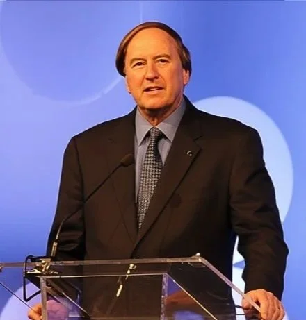 A man in a dark suit and tie speaking at a podium with microphones, standing against a blue background.