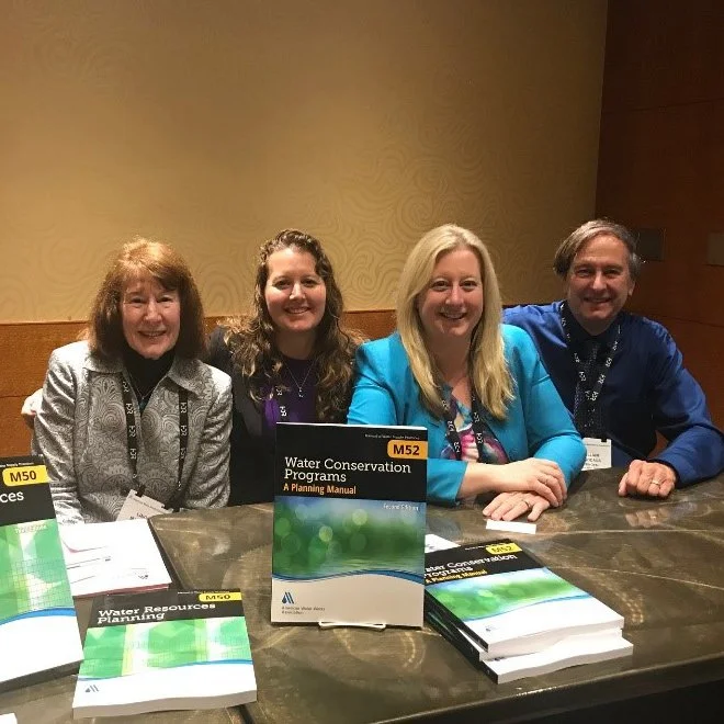 Four smiling people sitting at a table with water conservation program books, conference name tags, and lanyards in a conference room.