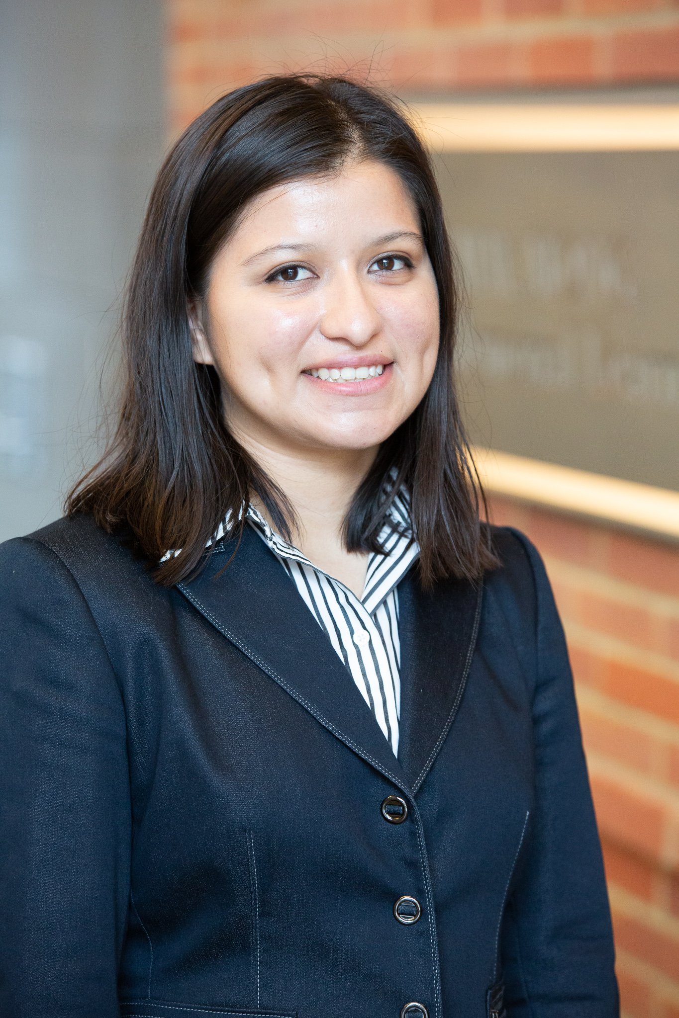 A professional woman with shoulder-length dark brown hair smiling at the camera, wearing a dark blazer over a striped collared shirt, with a blurred indoor background.