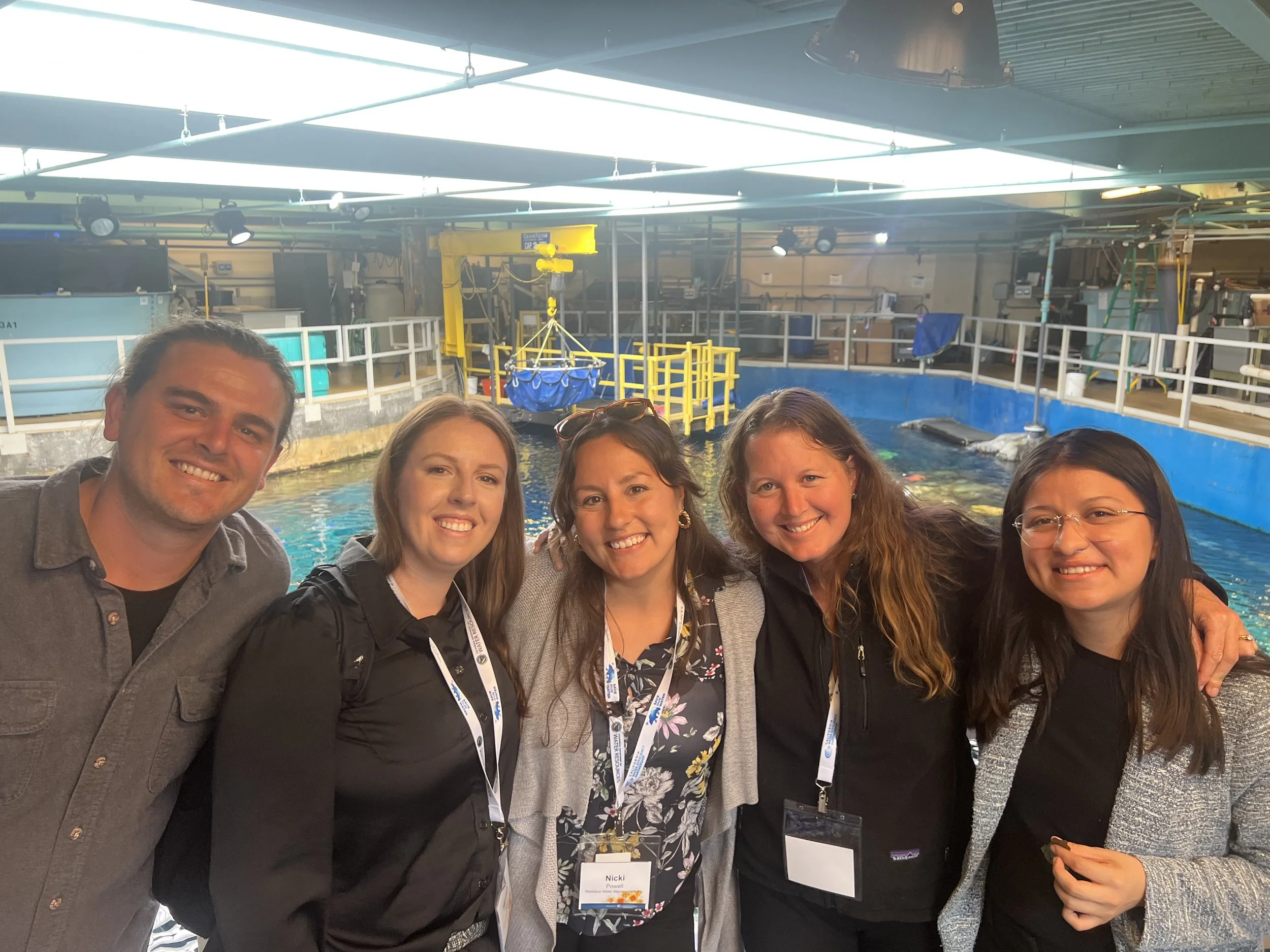 A group of five people posing together indoors near a large rectangular tank filled with water, with industrial equipment and lighting above.
