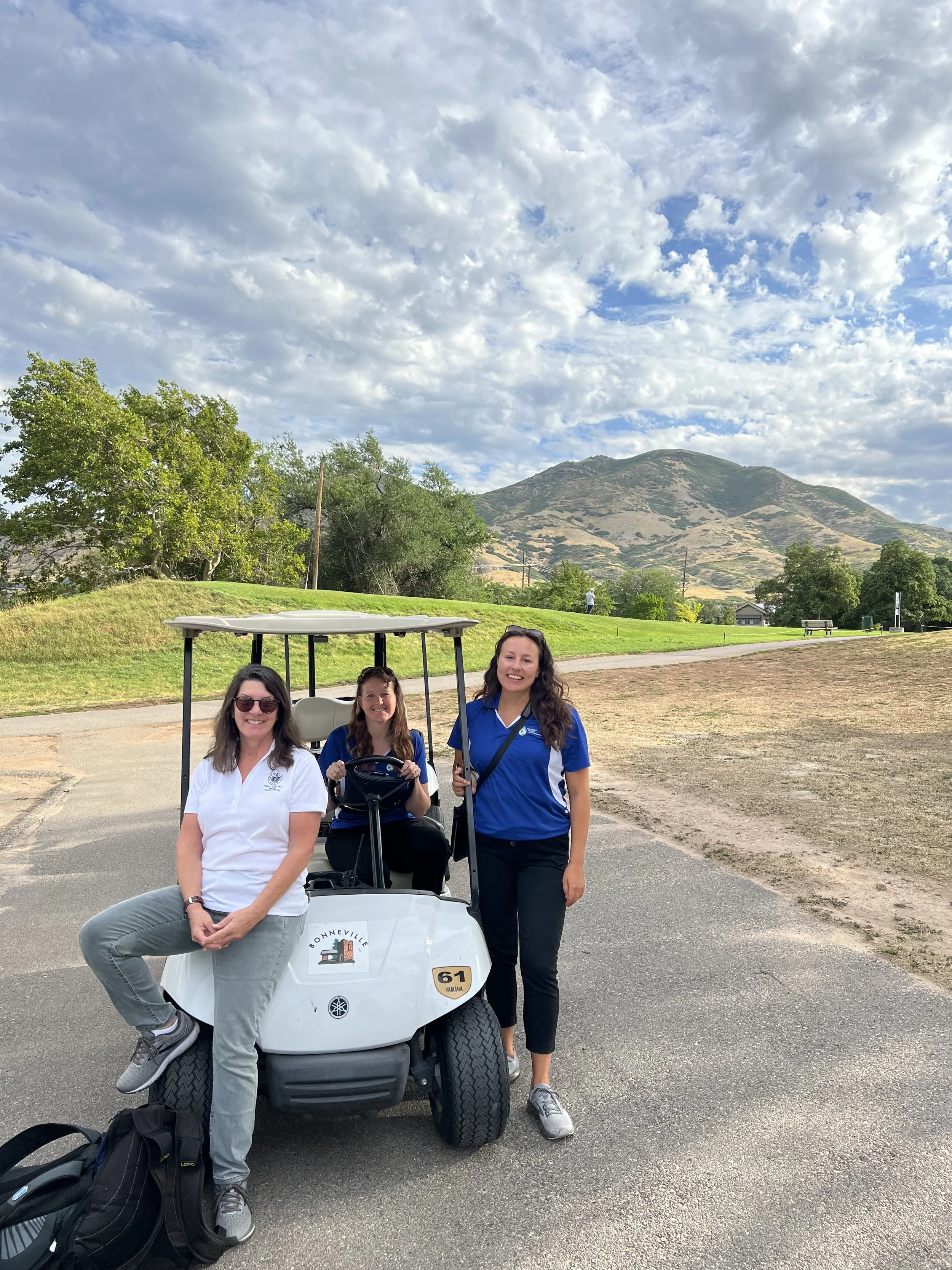 Three women at a golf course, one sitting on the golf cart, one standing beside it, and one driving it. The women are smiling, with a mountain and a partly cloudy sky in the background.